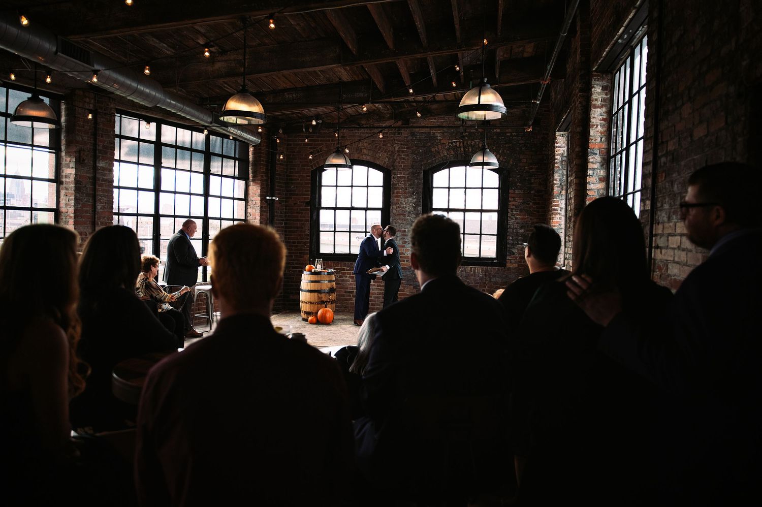 Silhouettes of people gathered inside a large industrial warehouse space with sunlight streaming through windows.