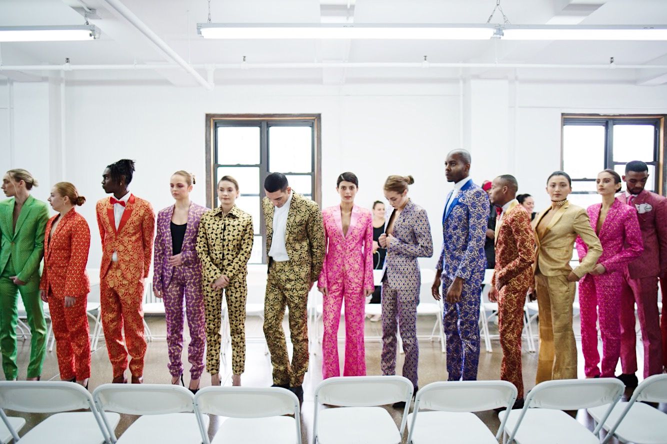 Models walk in a line showcasing colorful fashion designs against a bright white studio backdrop during a runway show.