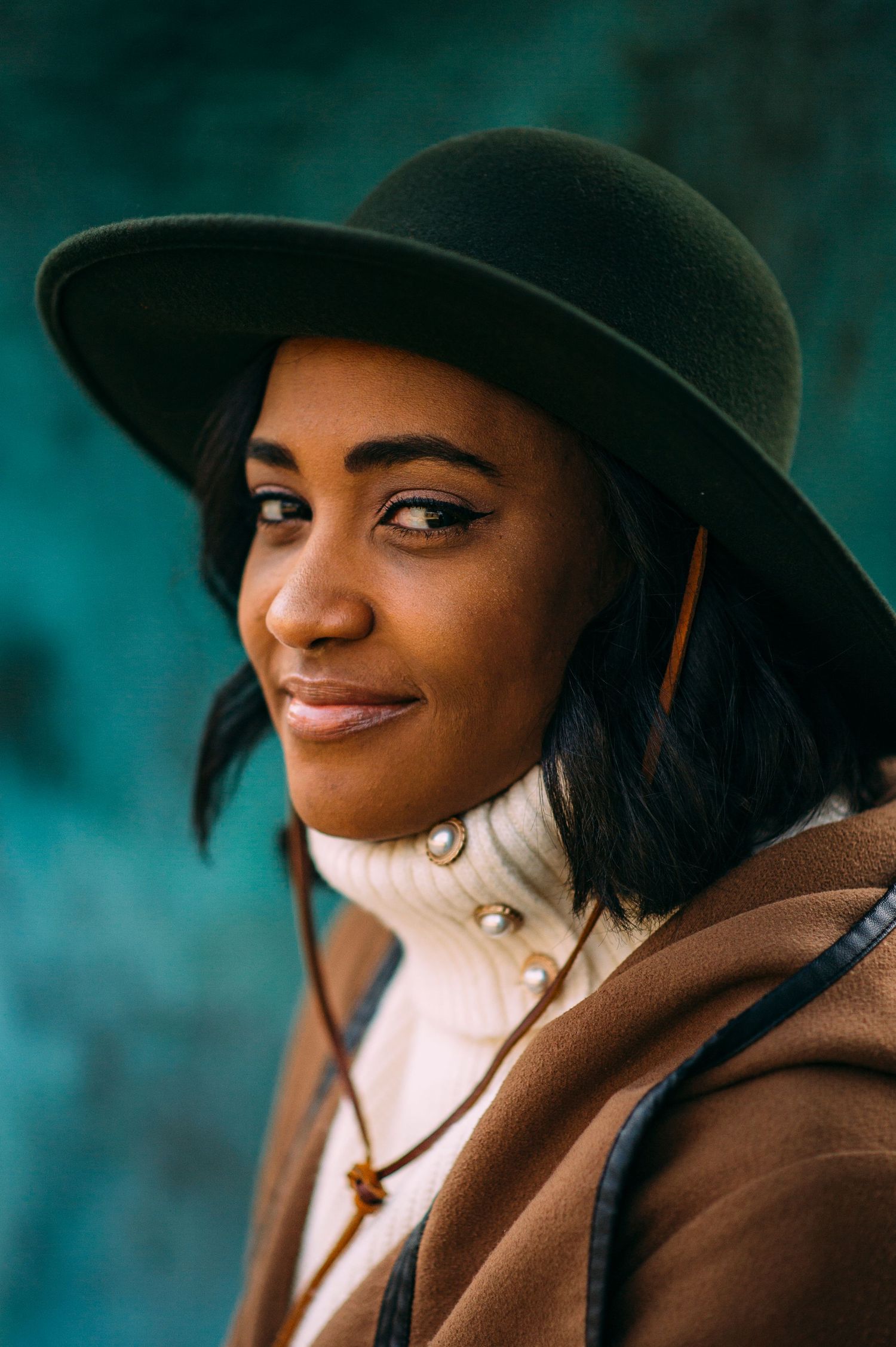 Stylish portrait wearing a wide-brimmed hat and brown coat against a teal background.