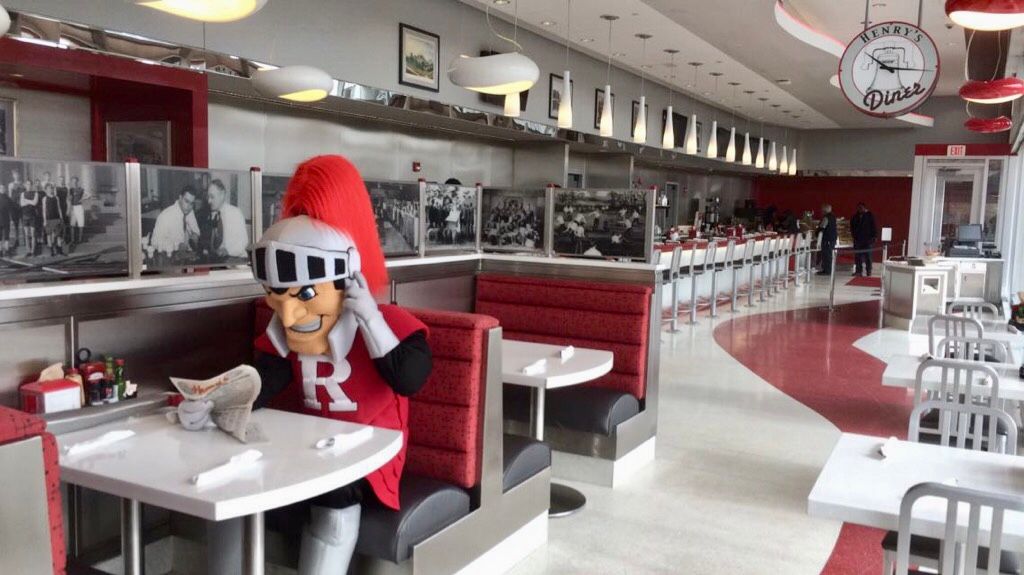 A panoramic view of a modern diner interior with red and chrome accents, featuring counter seating and vintage photographs.