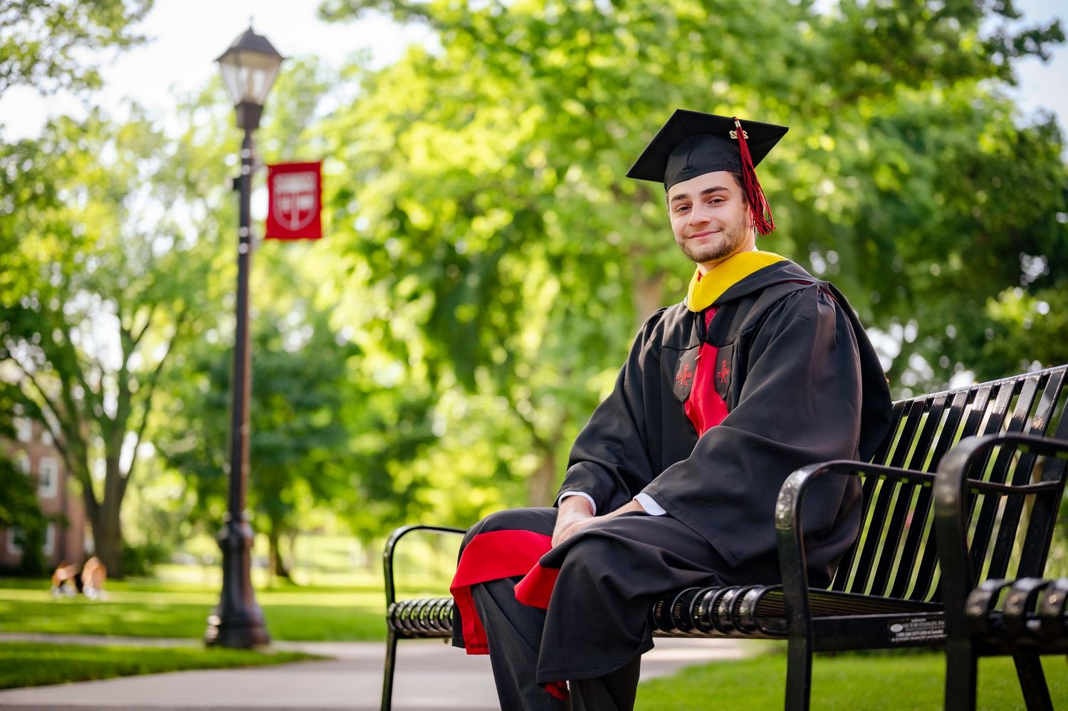 Graduate in black cap and gown sitting on bench in park setting.