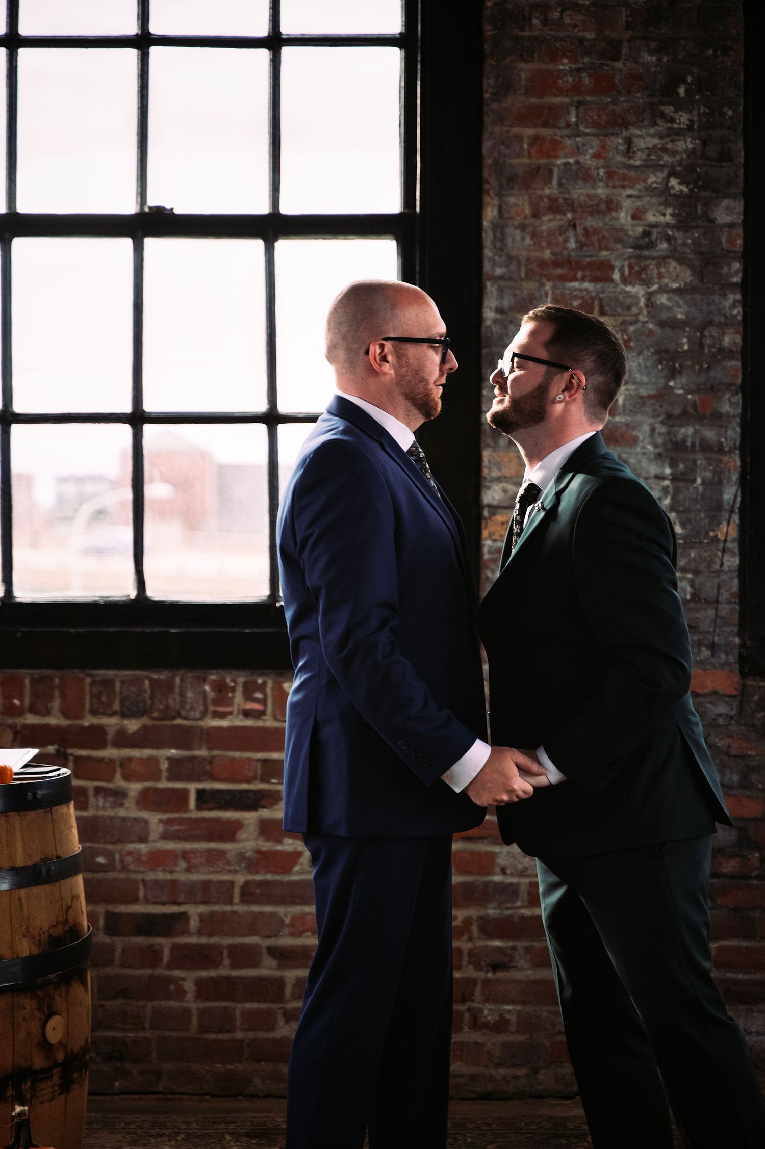 Two men in navy suits embrace in a rustic brick venue with natural window lighting creating a dramatic silhouette effect.