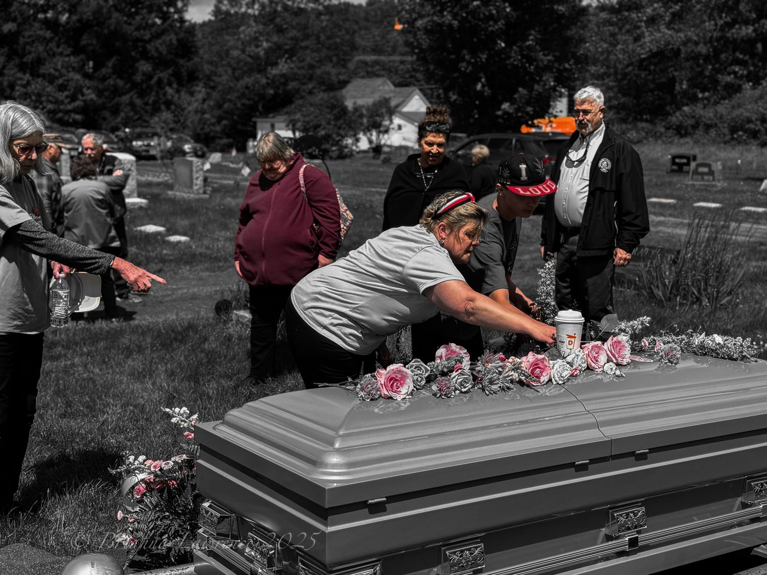 Black and white photo showing a funeral service with  flowers, and mourners paying their respects.