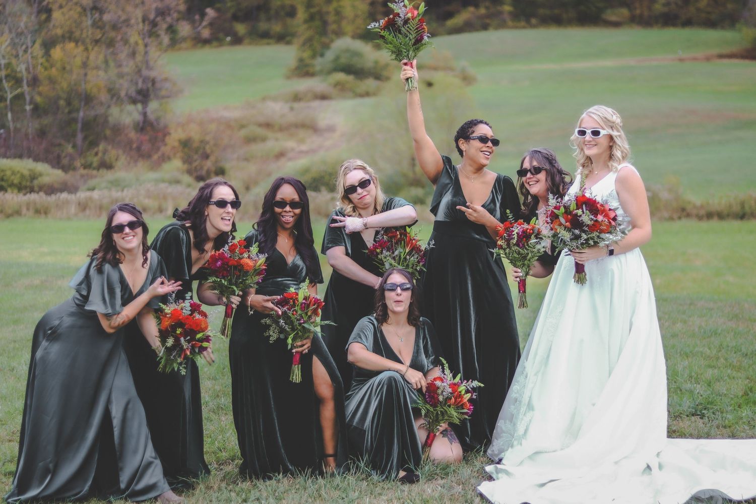 Wedding party poses together outdoors on a grassy hill during autumn with bridesmaids in green and colorful bouquets.