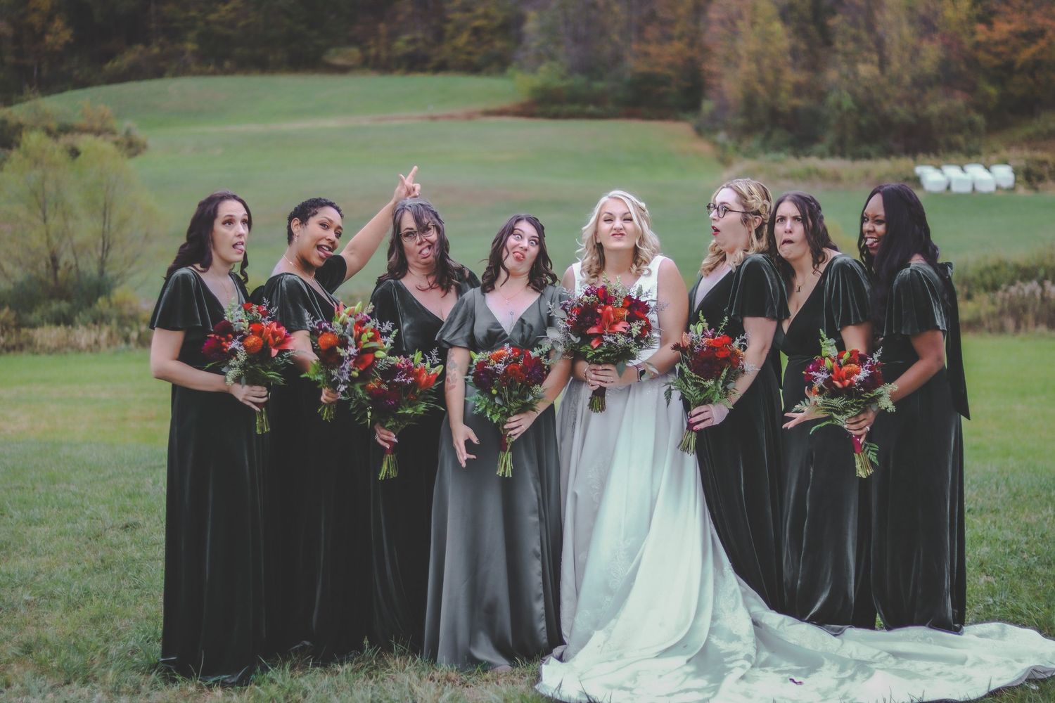 Wedding party in green dresses and autumn colors posing on a grassy hill.