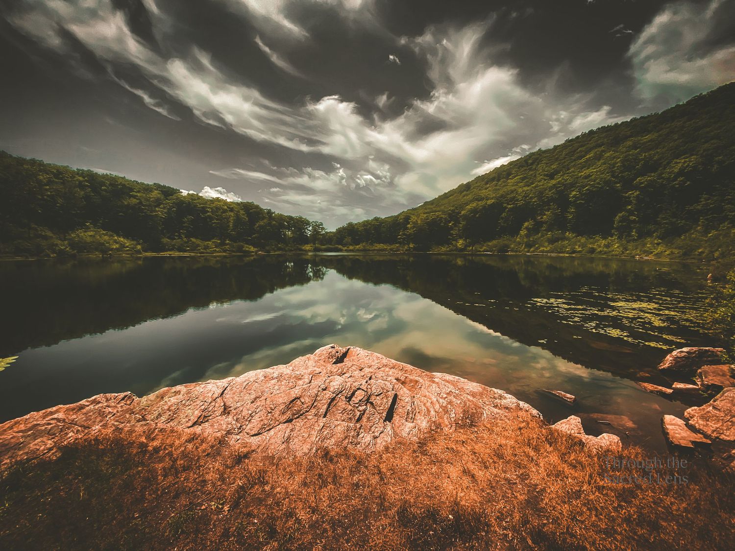 Dramatic mountain landscape with reddish rocks and a lake under sweeping clouds.