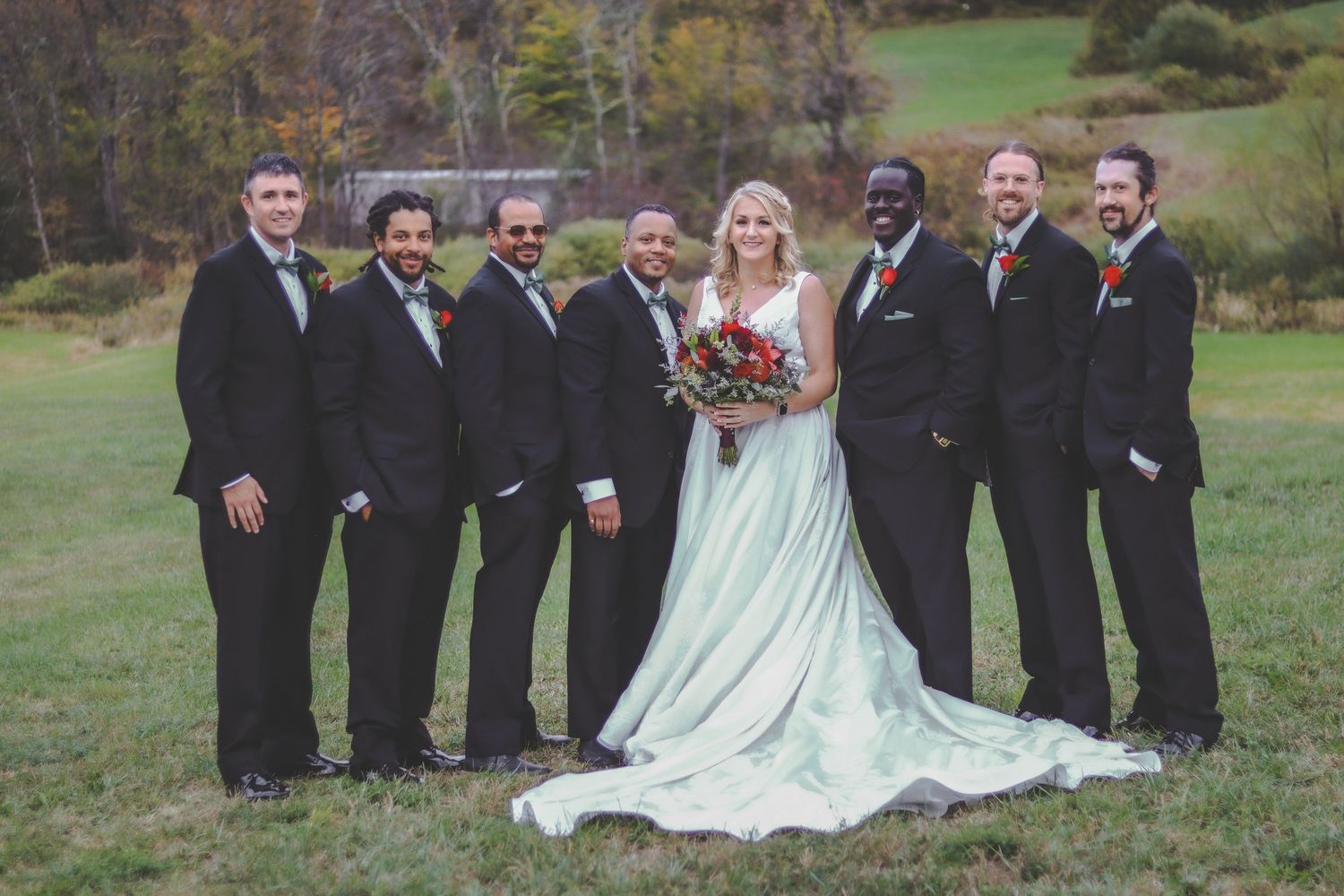Wedding party stands in formation on green lawn with bridesmaids in black dresses and groomsmen in dark suits.