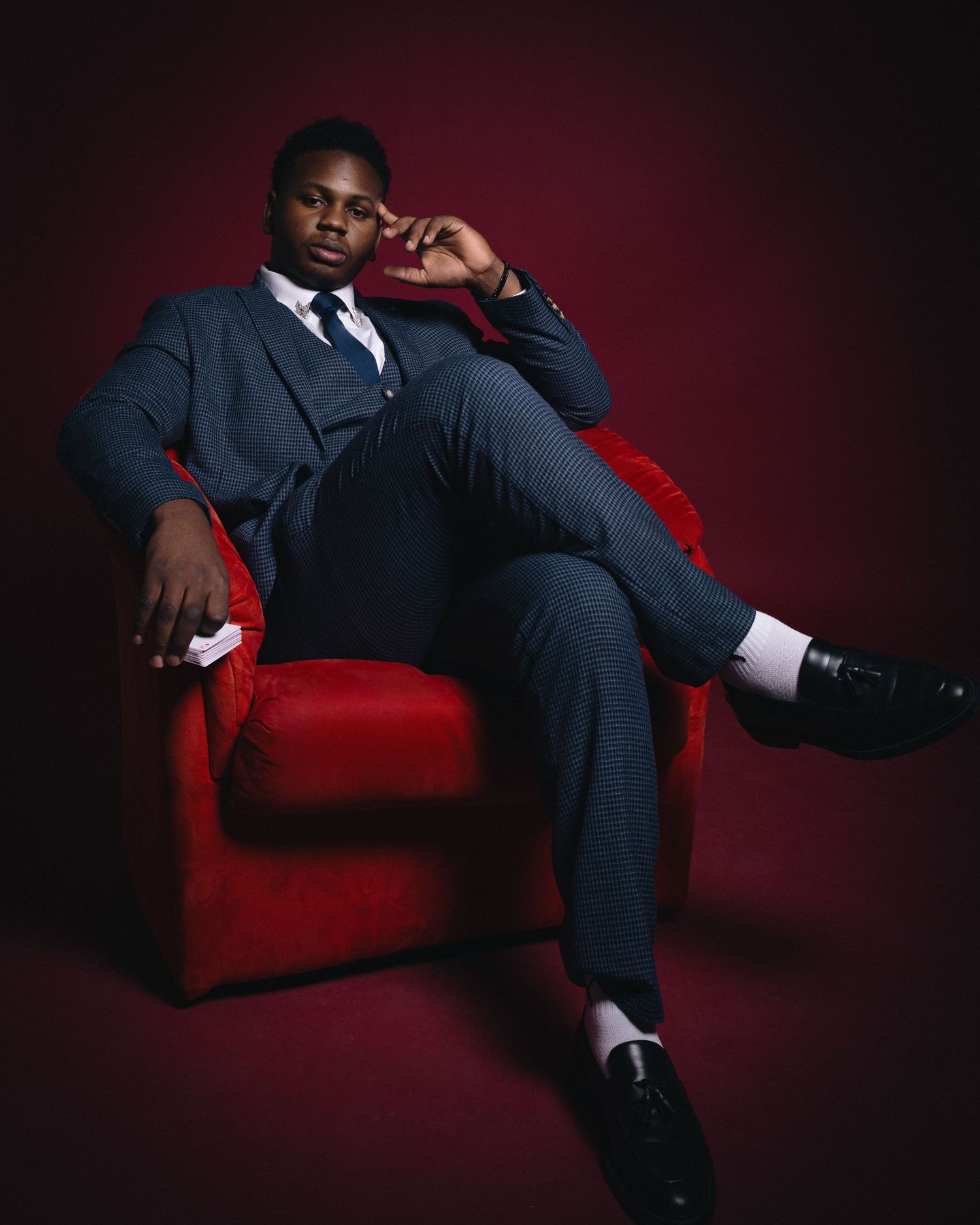 Man in a blue suit seated in a red chair against a dark red backdrop.