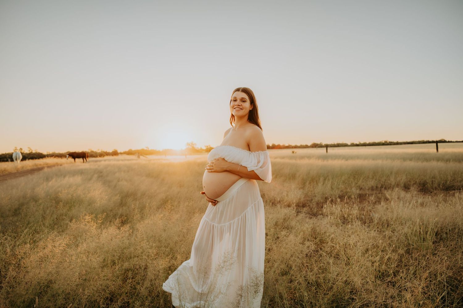 Pregnant woman in white lace dress holding baby bump at golden sunset in open field.