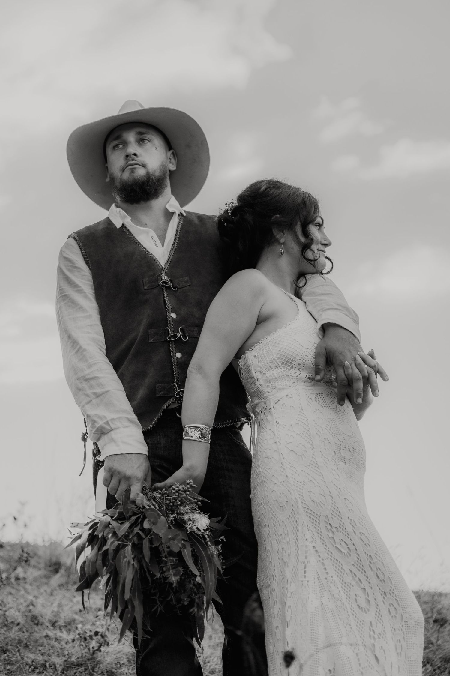 Black and white photo of a bearded groom in cowboy hat holding bouquet with bride in lace dress outdoors.