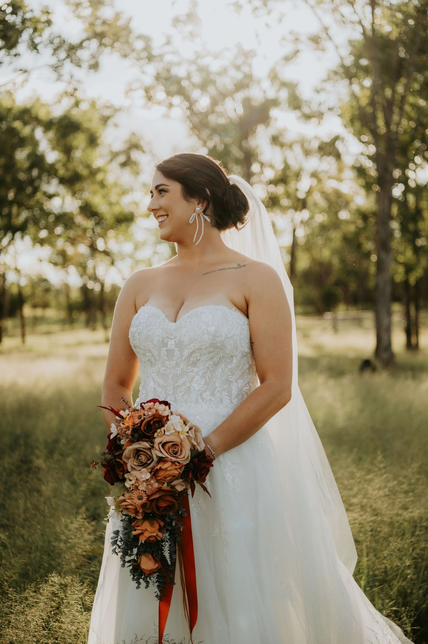 Bride in strapless lace wedding gown holding autumn bouquet of roses stands in sunlit meadow with trees.