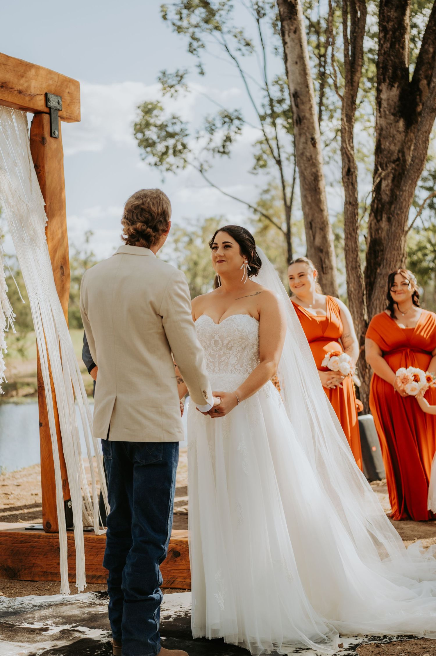Bride in white gown holds hands with groom at outdoor wedding ceremony with bridesmaids in orange dresses.