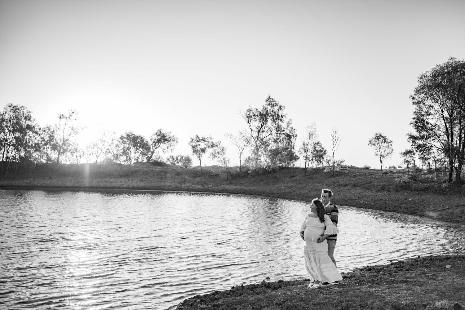 A panoramic black and white landscape shows a serene lakeside scene with trees reflecting on calm waters at sunset.