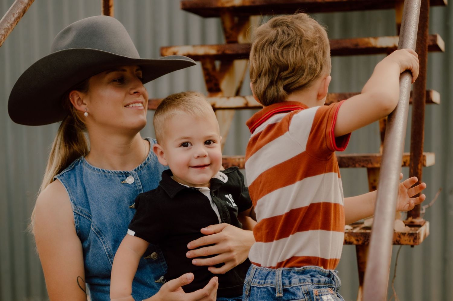 Woman in cowboy hat holds smiling baby in black outfit while boy in striped shirt climbs rustic metal ladder outdoors.