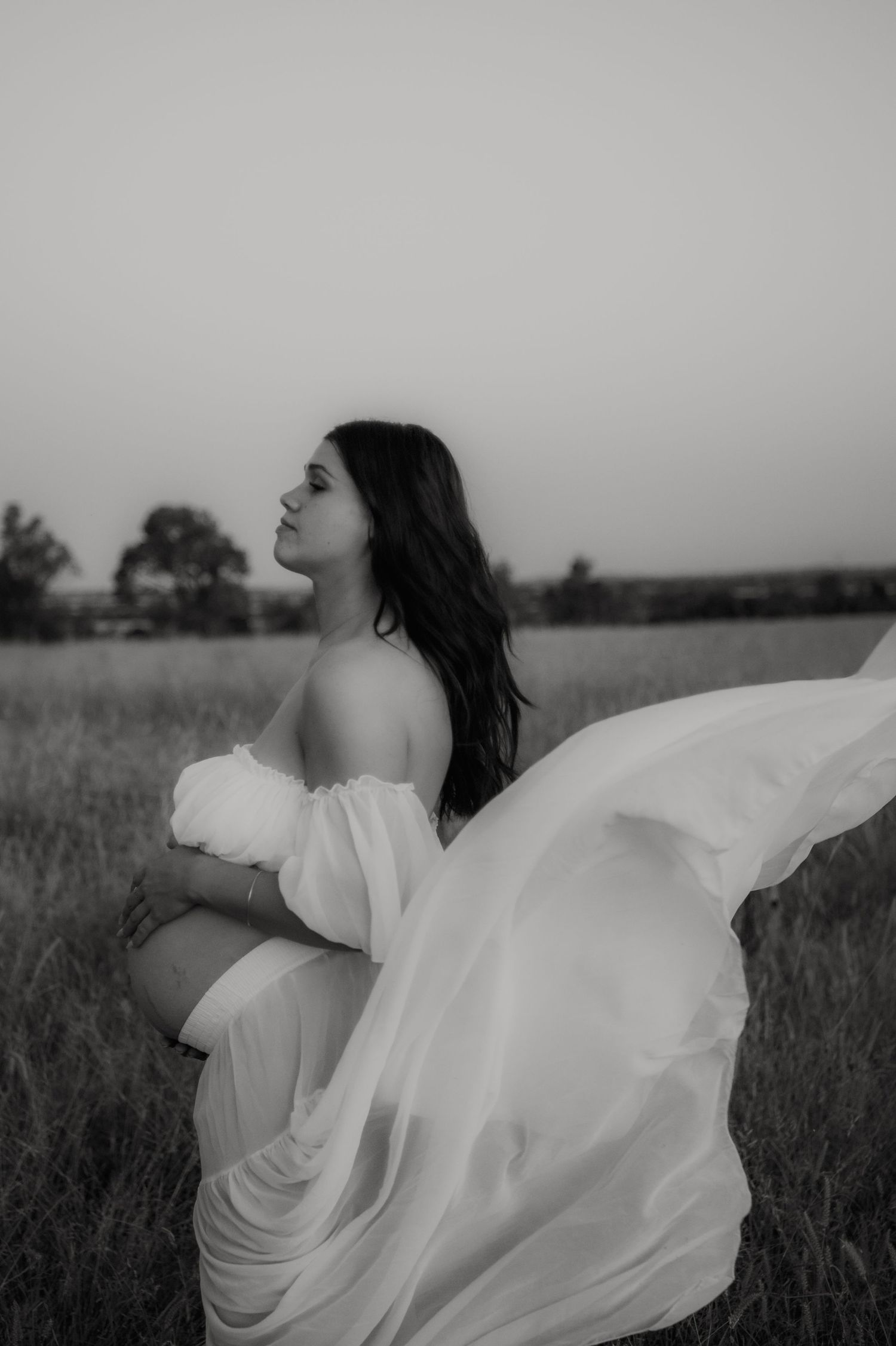 Black and white photo of a pregnant woman in a flowing white dress standing in a field at dusk.