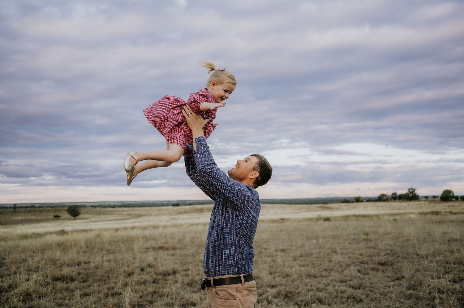 Father joyfully lifting his young daughter high in the air in an open grassy field under a cloudy sky.