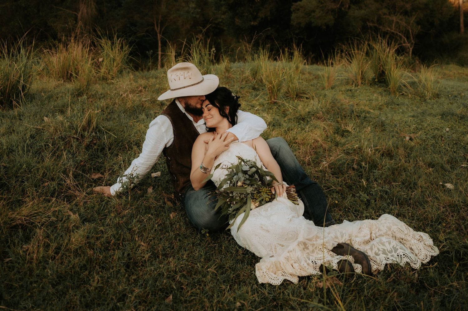 Romantic couple sitting in lush green field, bride in lace dress with bouquet, groom in cowboy hat, sharing tender moment.