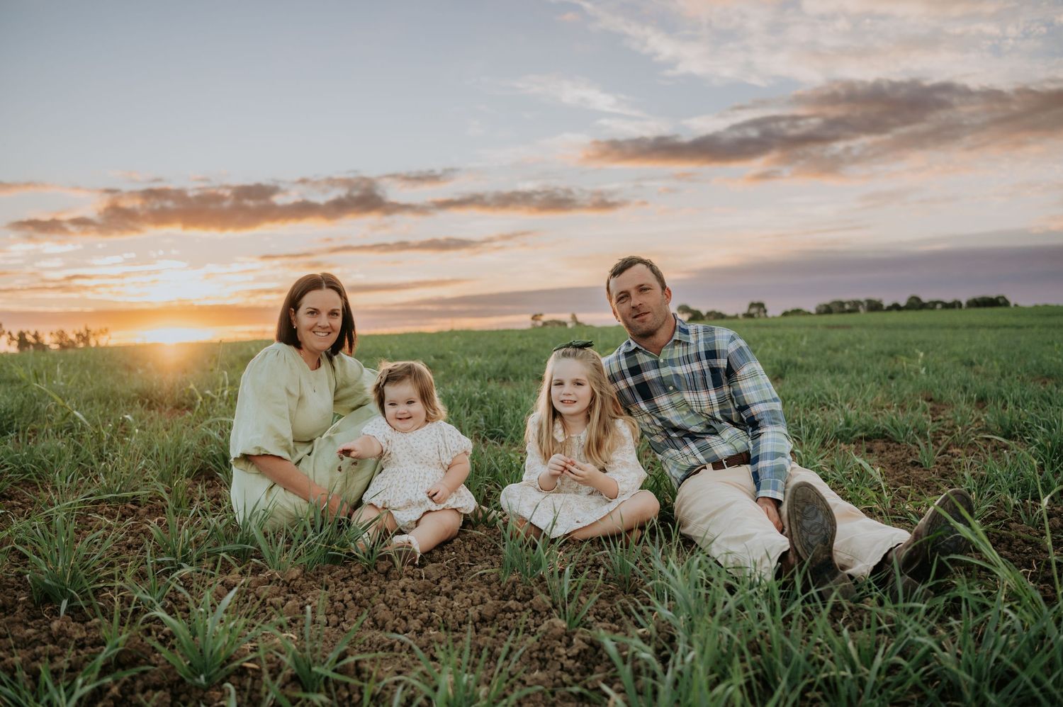 Family of four sitting in a green field at sunset, smiling, with a colorful sky backdrop.
