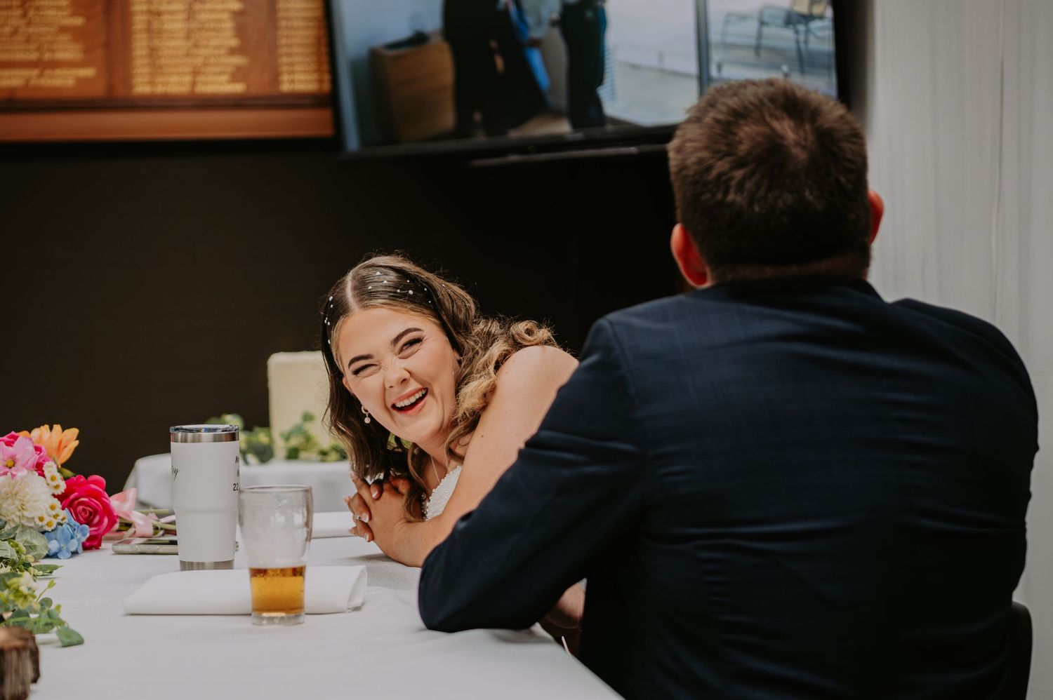 Bride laughing joyfully at wedding reception table with colorful flowers and drinks nearby.