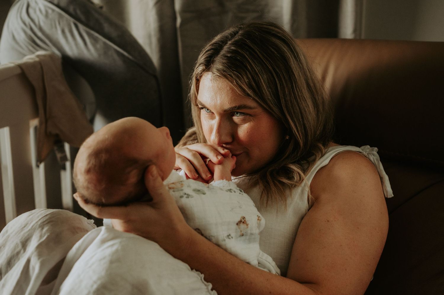 A mother tenderly gazes at her newborn baby while sitting in a leather chair, wrapped in a white blanket.
