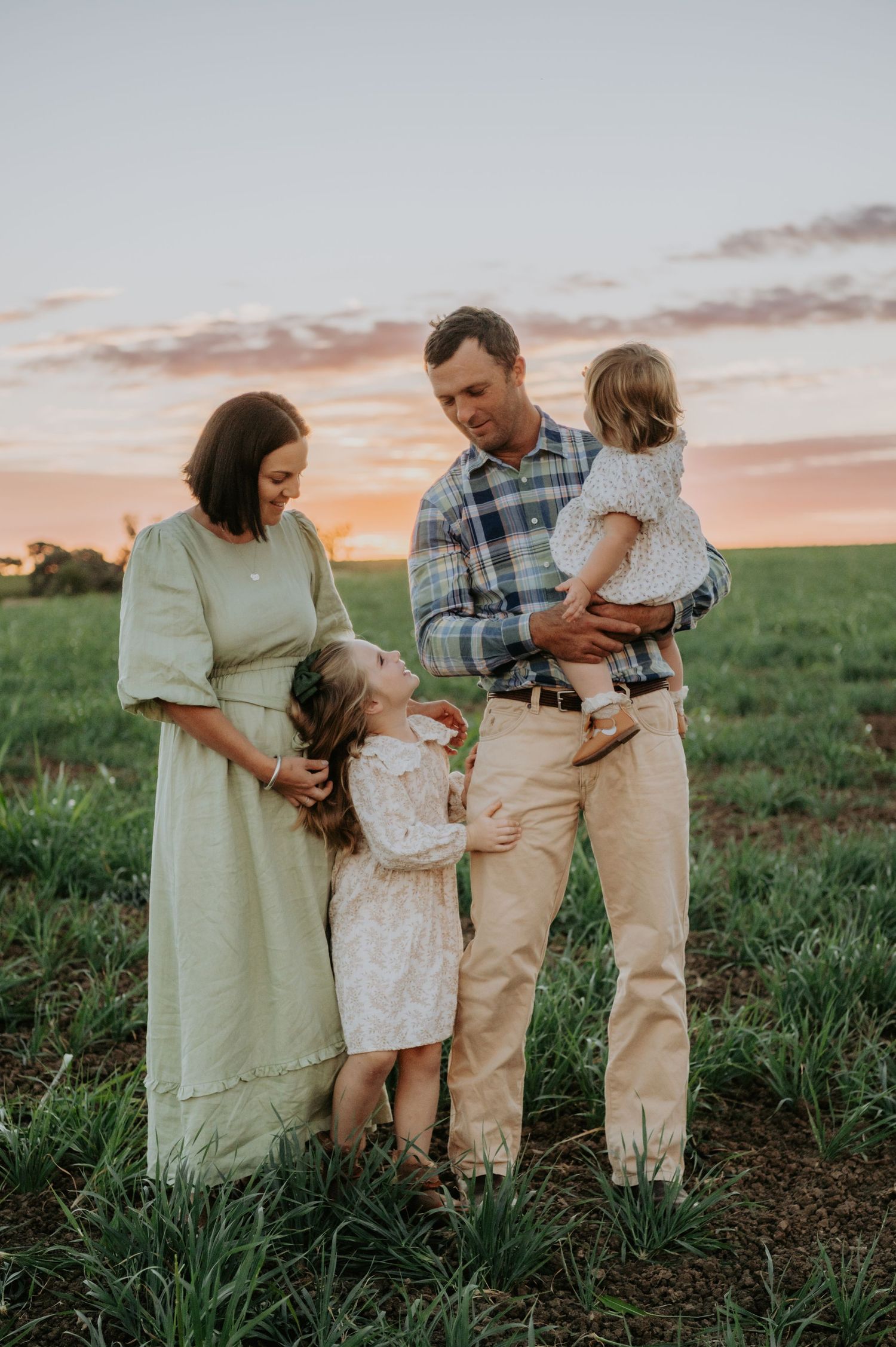 Family of four posing in a green field at sunset, parents with two young children in soft natural light.