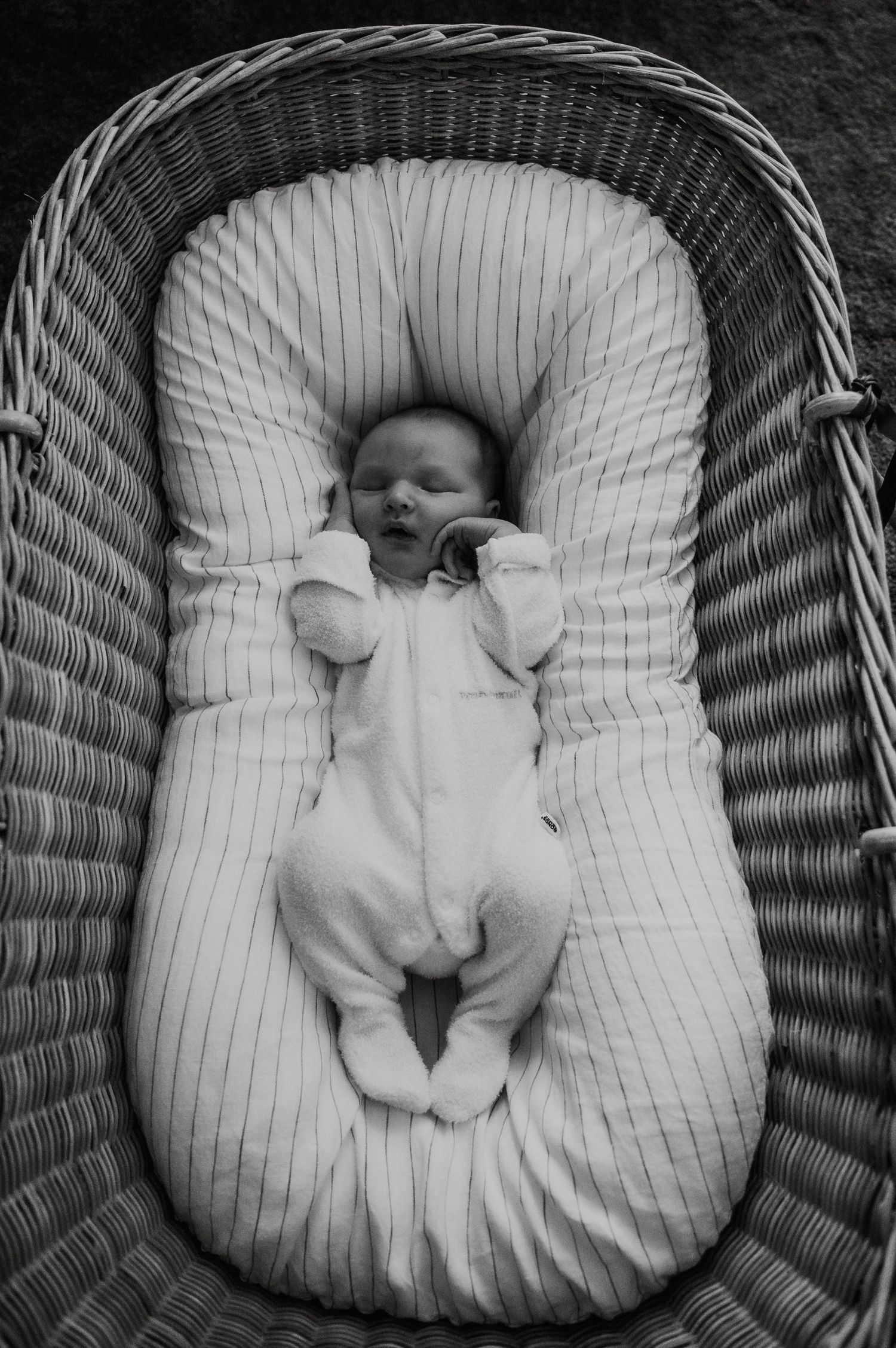 Black and white image of sleeping baby in white onesie lying in wicker bassinet.