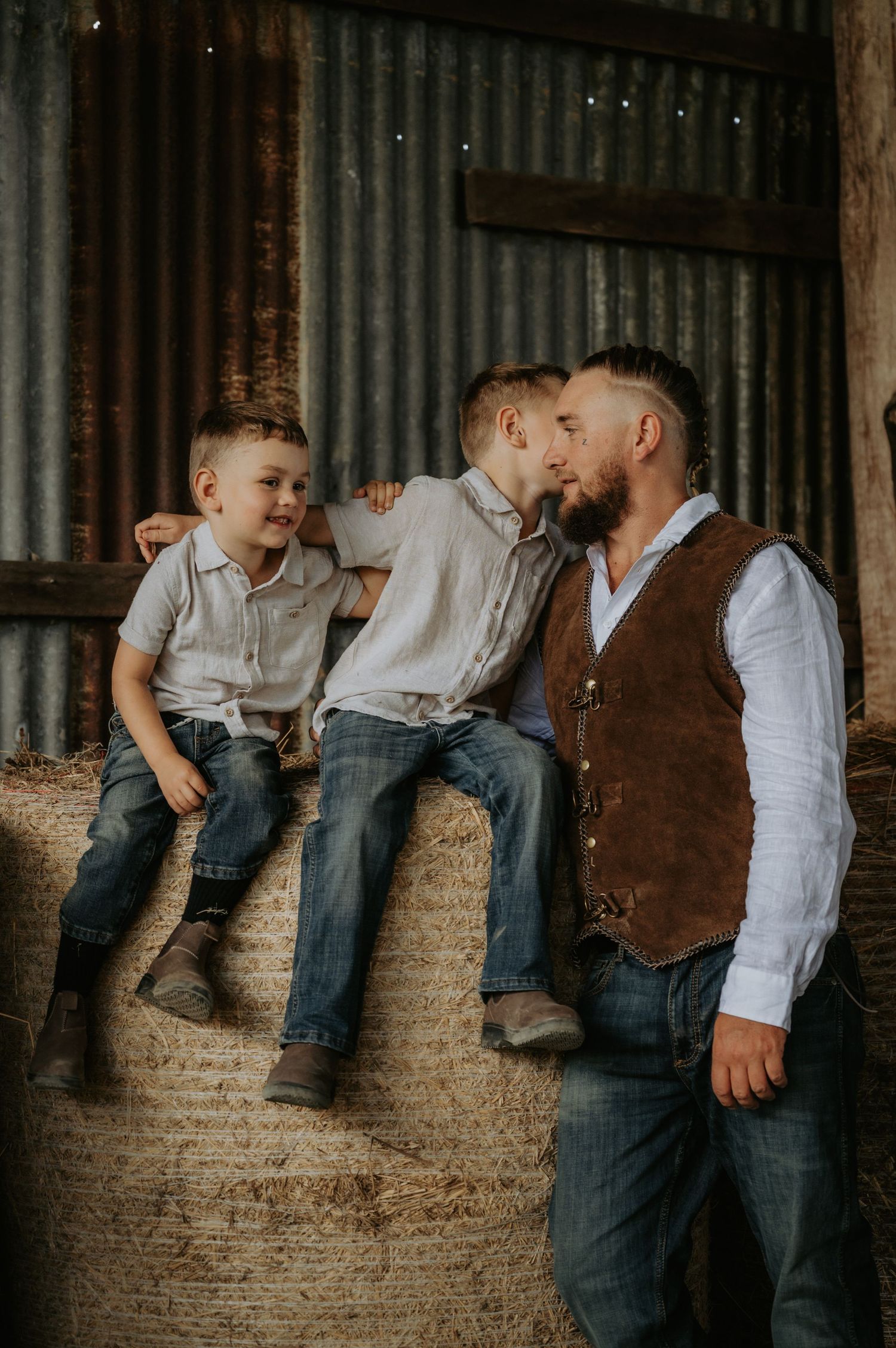 A father and two young boys share a tender moment on hay bales inside a rustic barn, dressed in casual western attire.