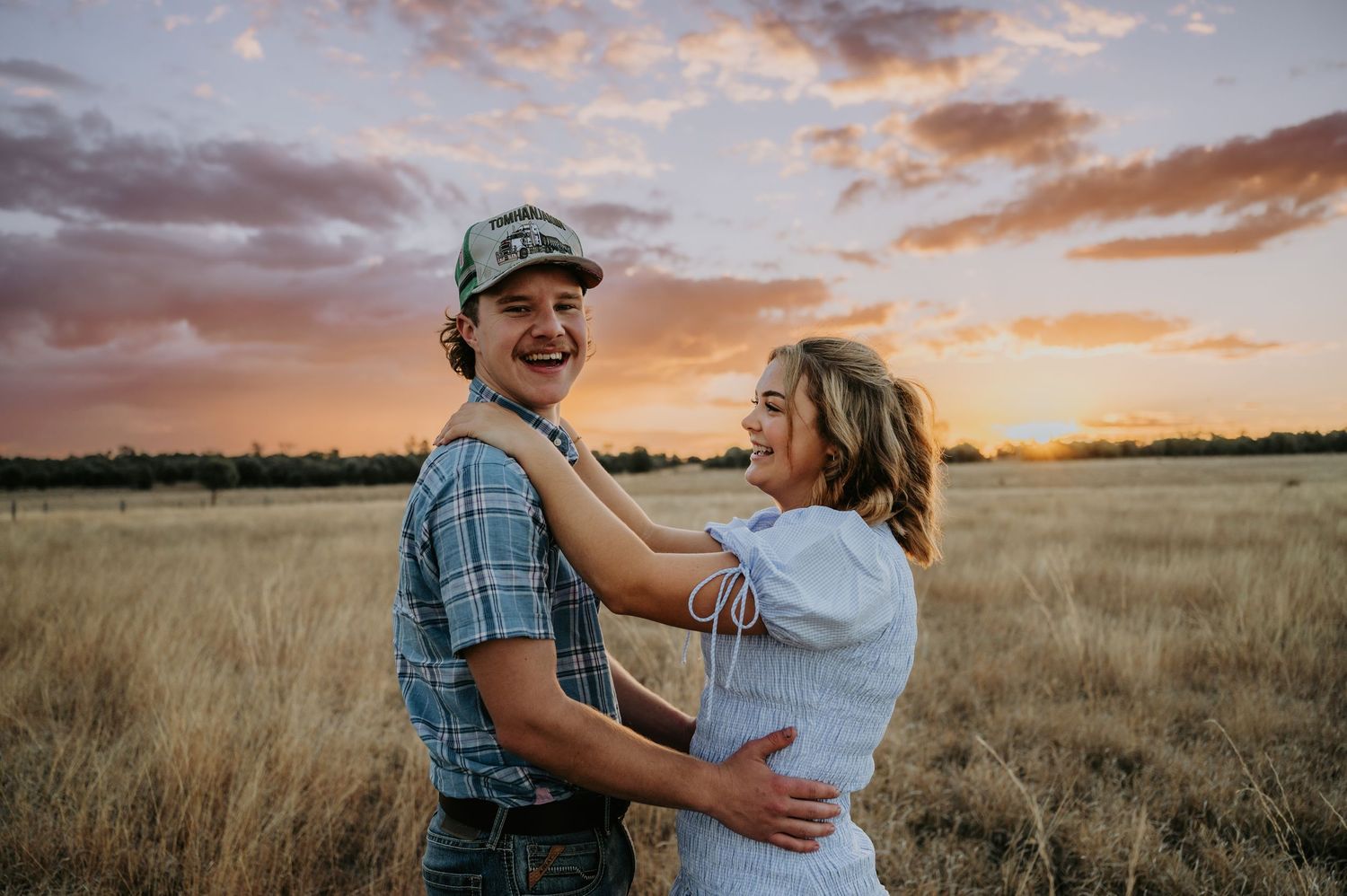 Couple laughing and embracing in golden field at sunset, man in plaid shirt and cap, woman in white dress.