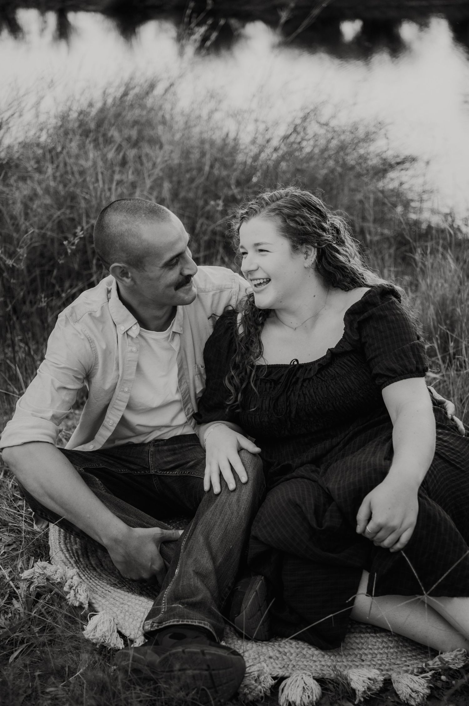 Black and white photo of a laughing couple sitting on a blanket in a grassy field near water.