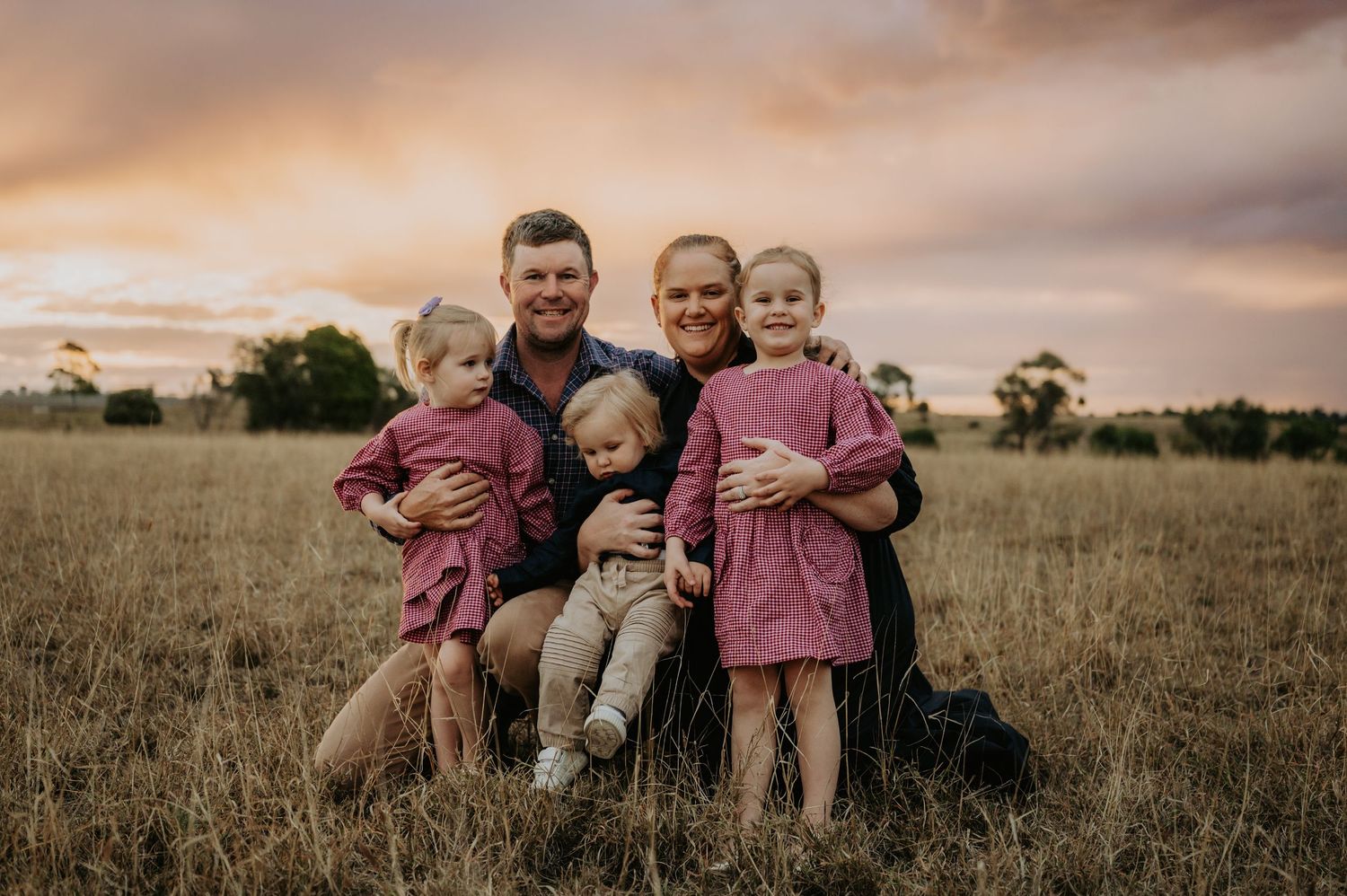 Family of five posing in golden field at sunset, parents holding three young children in matching pink outfits.