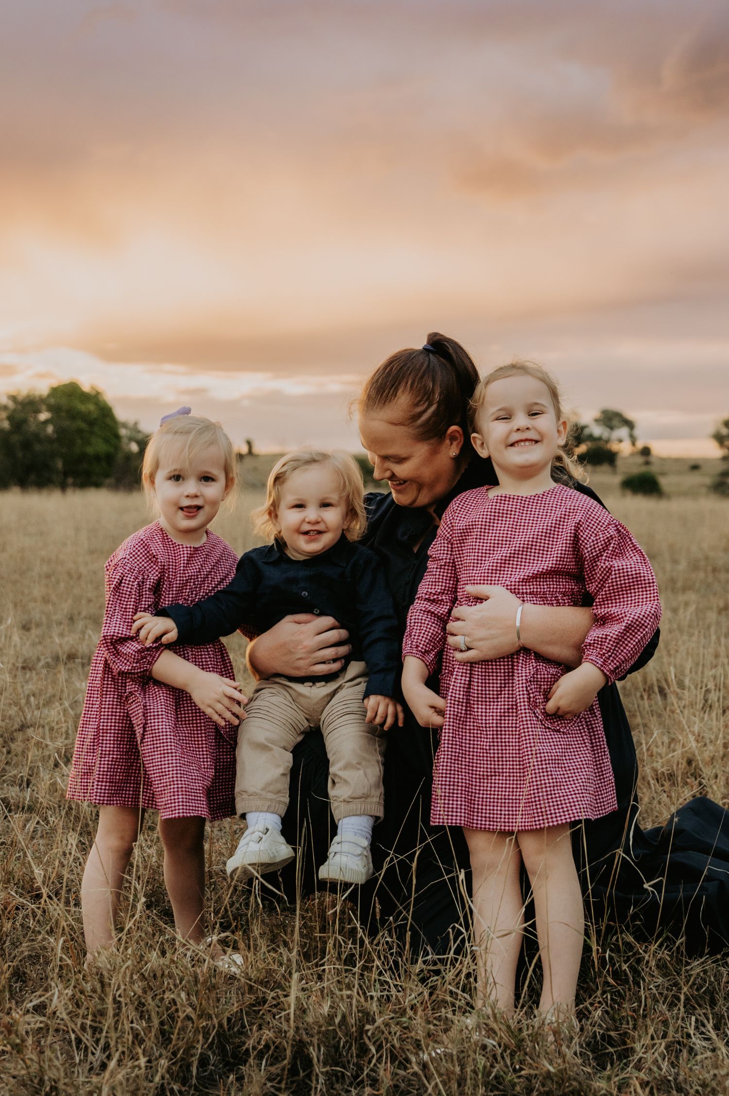 A mother kneels in a golden field at sunset, smiling with her three young children in matching pink gingham dresses.