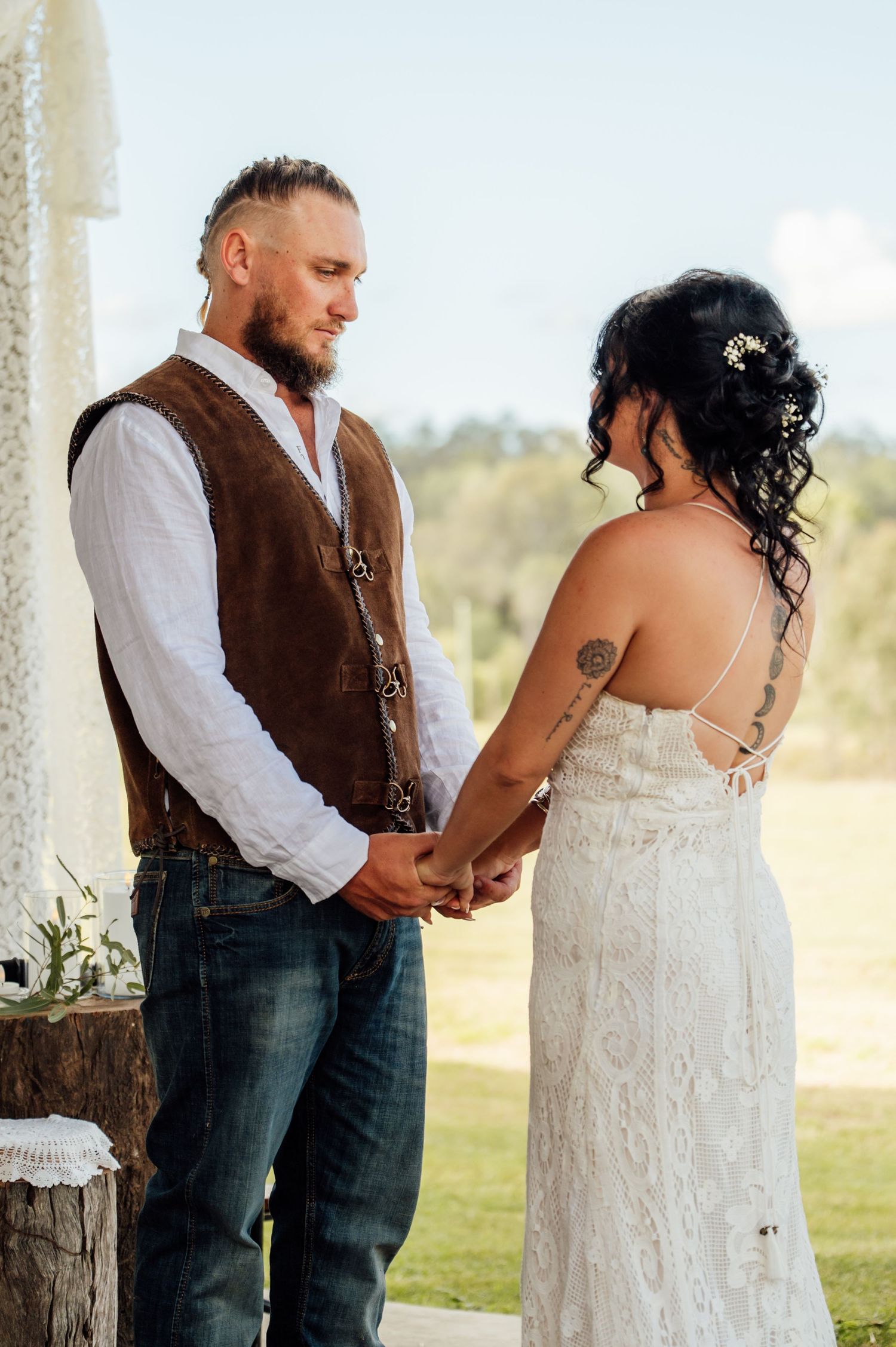 Couple holding hands at outdoor boho wedding ceremony, groom in brown vest and jeans, bride in lace dress with tattoos.