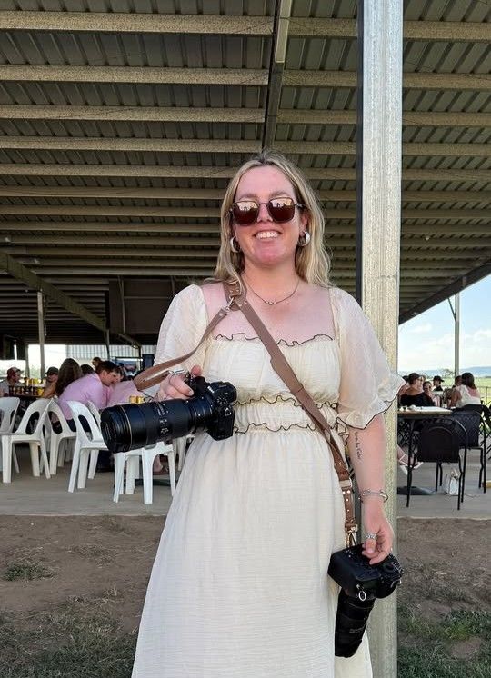 Professional photographer in white dress holding camera equipment under covered outdoor area.