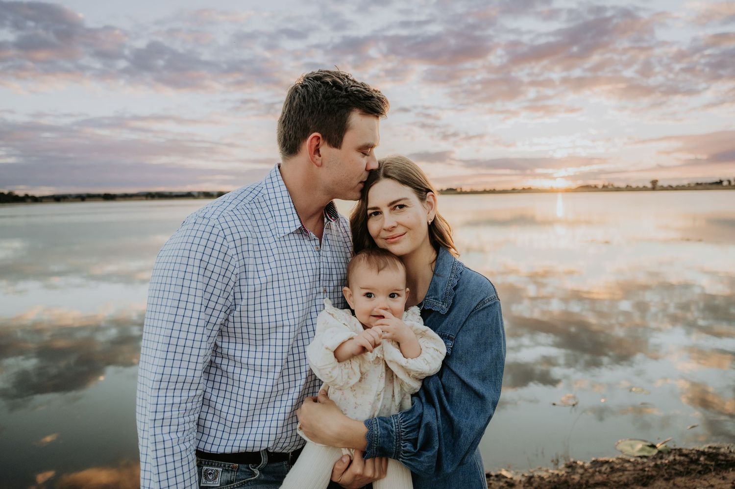 A young family poses by a lake at sunset, parents holding their baby girl in a warm, romantic outdoor portrait.