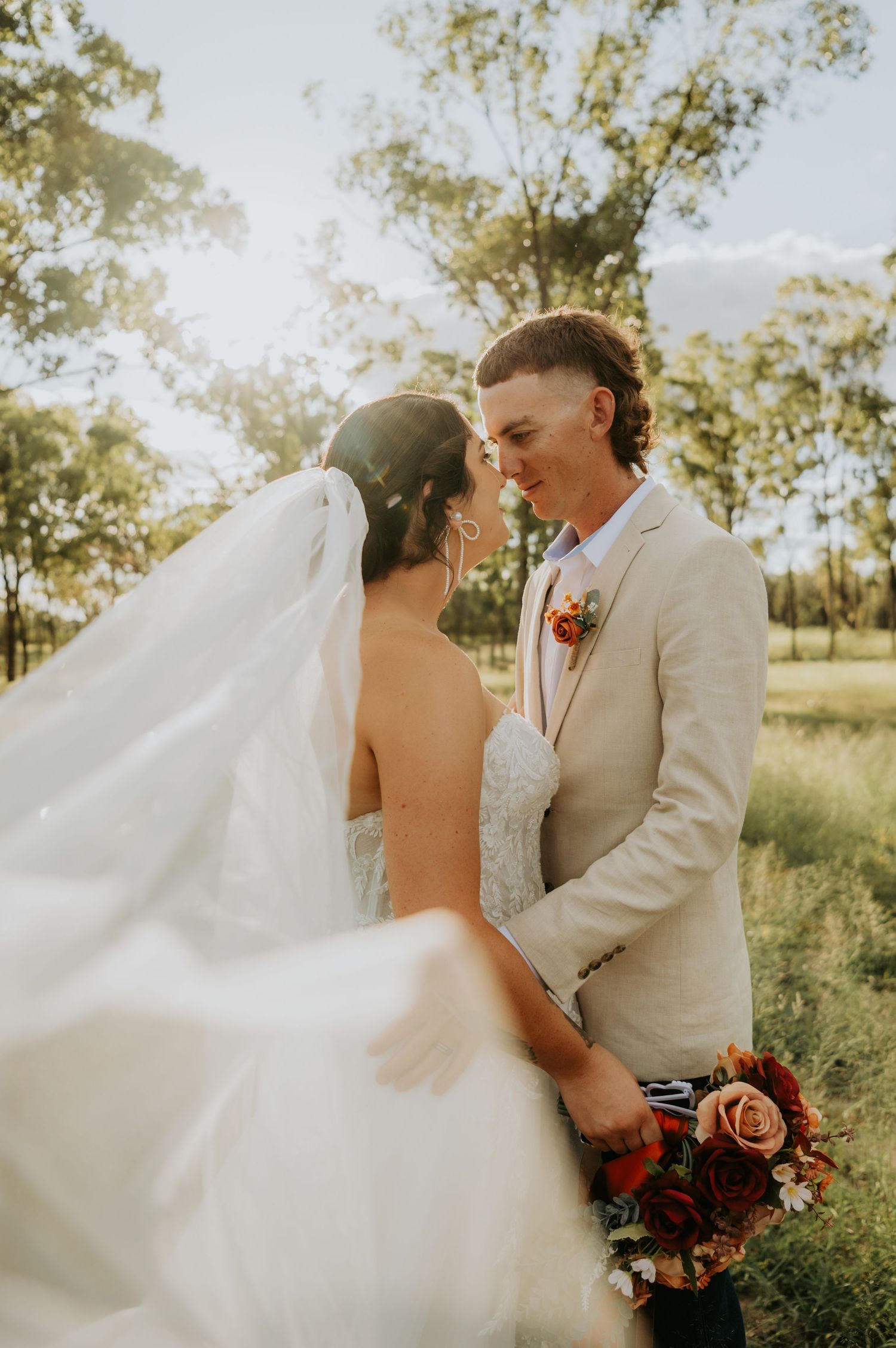 Bride and groom share a romantic moment outdoors, with flowing veil, tan suit, and autumn bouquet in a sunlit garden.
