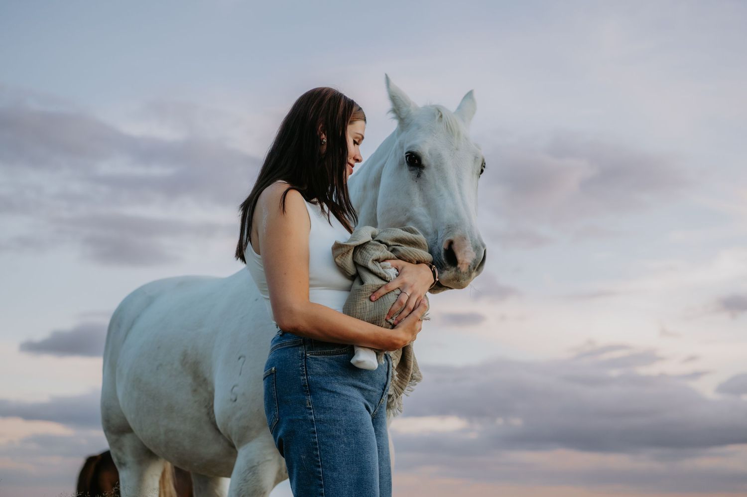 Young woman in jeans tenderly embracing a white horse against a pastel cloudy sky at dusk.