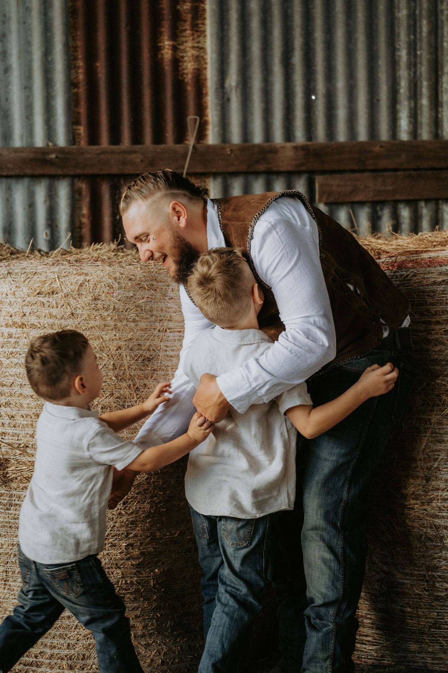 A father in a vest and white shirt laughs as two young boys playfully tug at him near hay bales in a rustic barn.