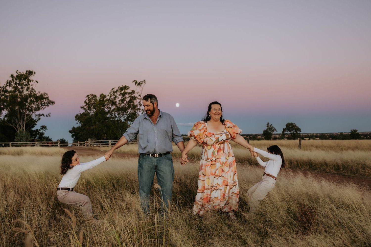 Family of four holding hands in a golden field at sunset with a full moon rising in the pastel sky.