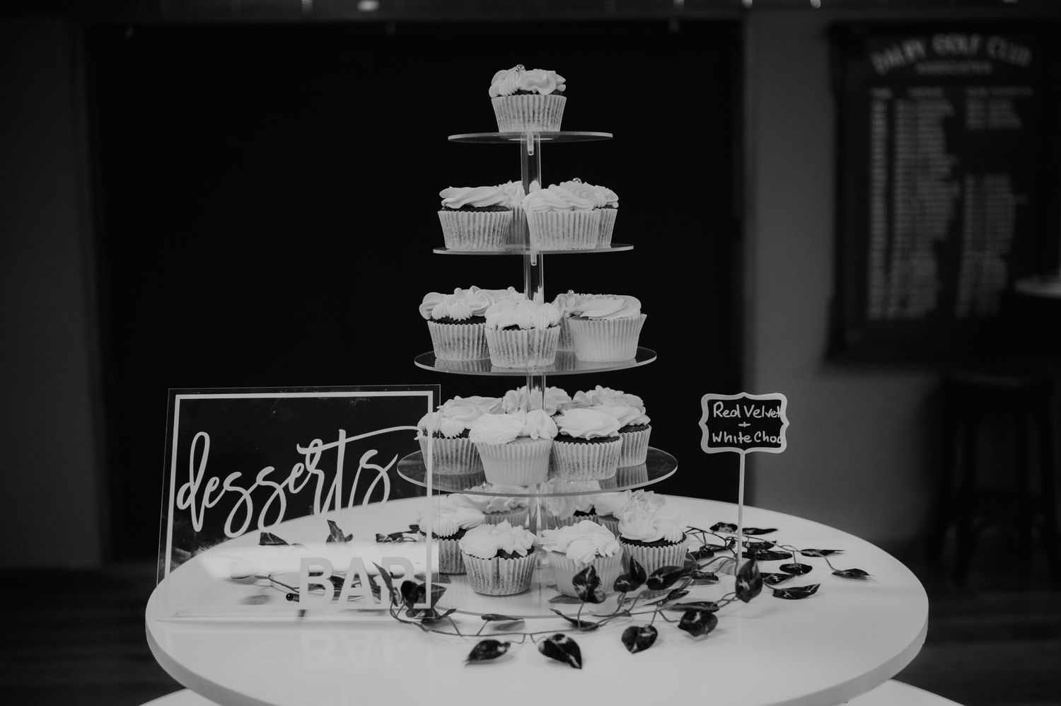 Black and white photo of a tiered cupcake tower display on a round table at a wedding dessert bar.