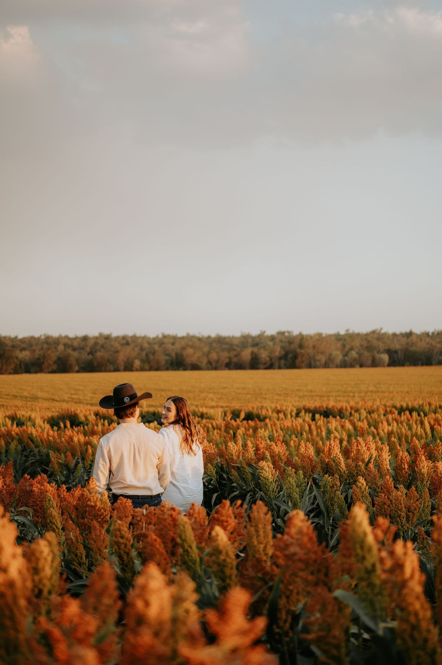 A couple walks through a golden wheat field at sunset with autumn trees in the background.