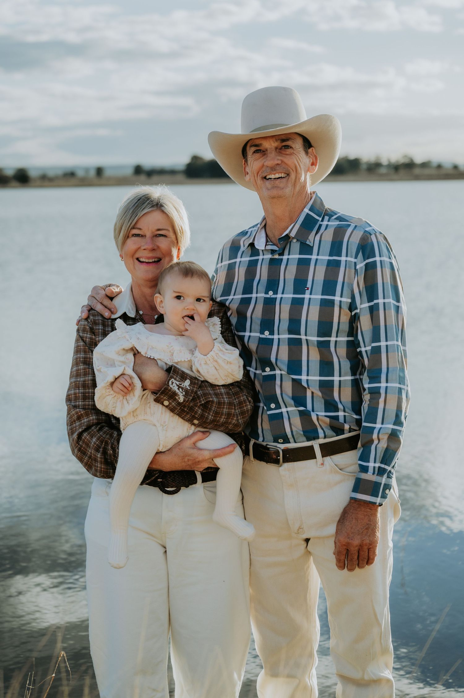 Smiling grandparents holding a happy baby outdoors near a lake, man wearing a cowboy hat and plaid shirt.