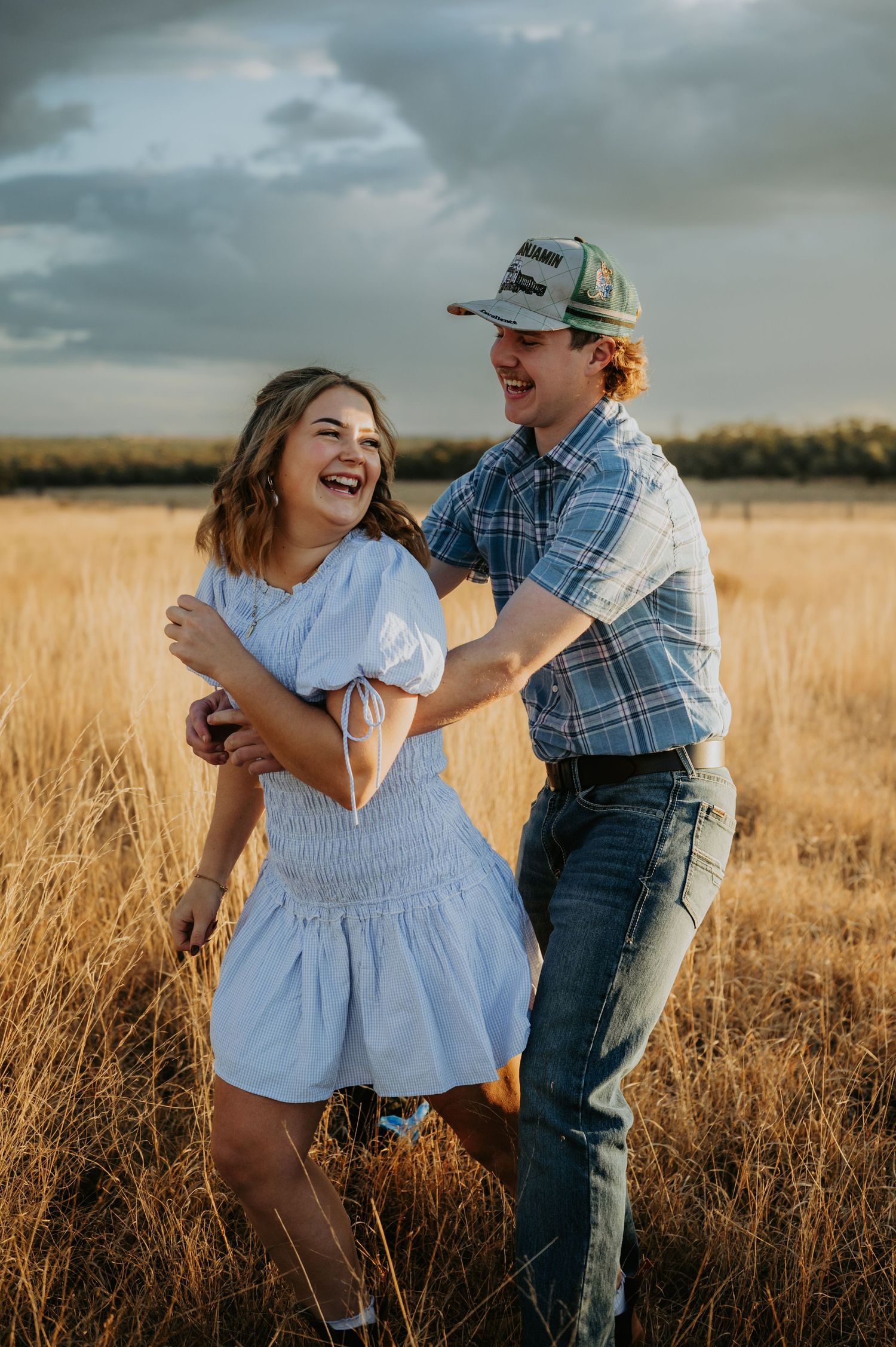 A laughing couple embraces in a golden wheat field, woman in white dress, man in plaid shirt and cap, under cloudy sky.
