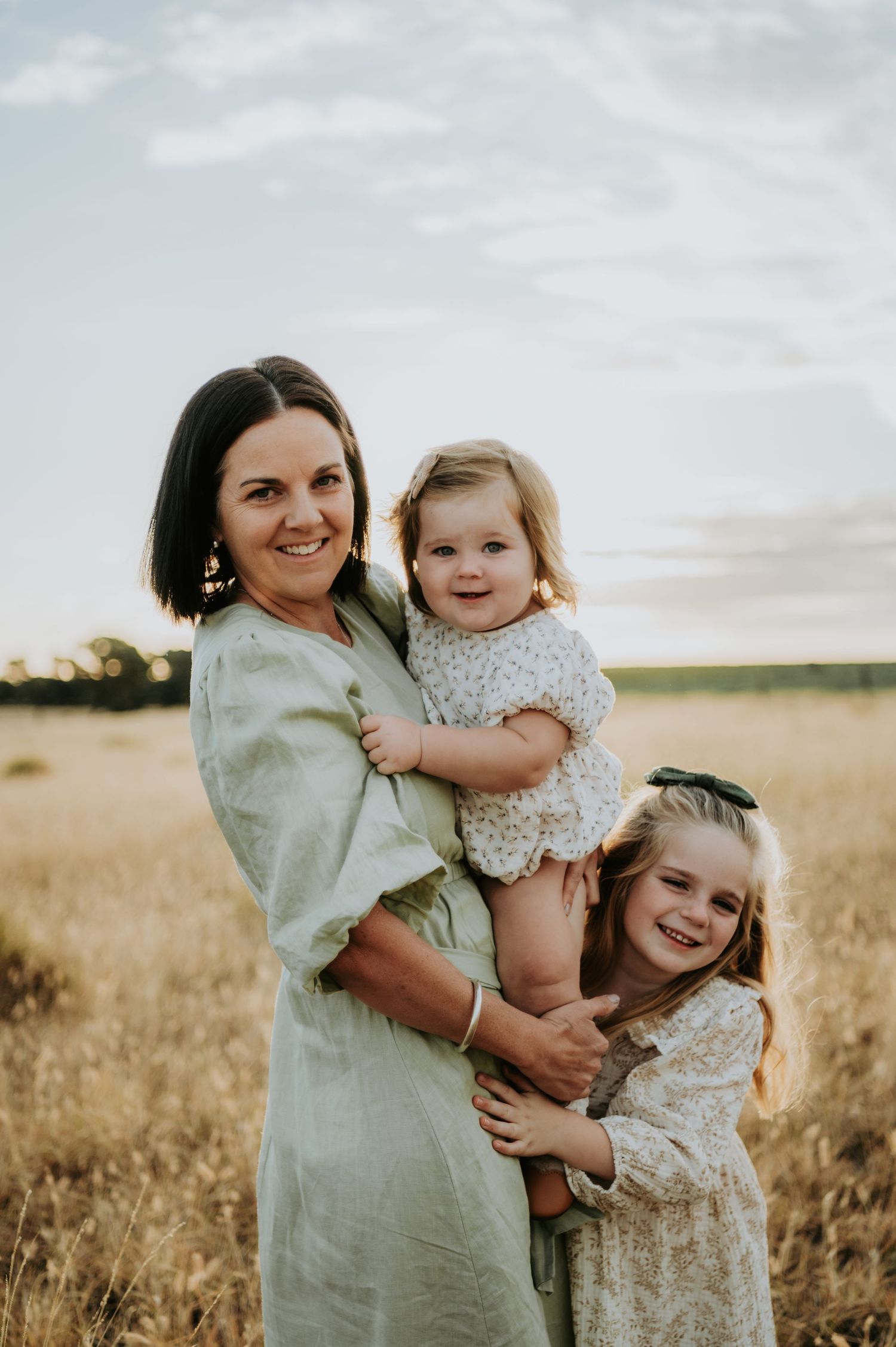 A smiling mother holds two young daughters in a golden field at sunset, wearing soft floral and linen dresses.