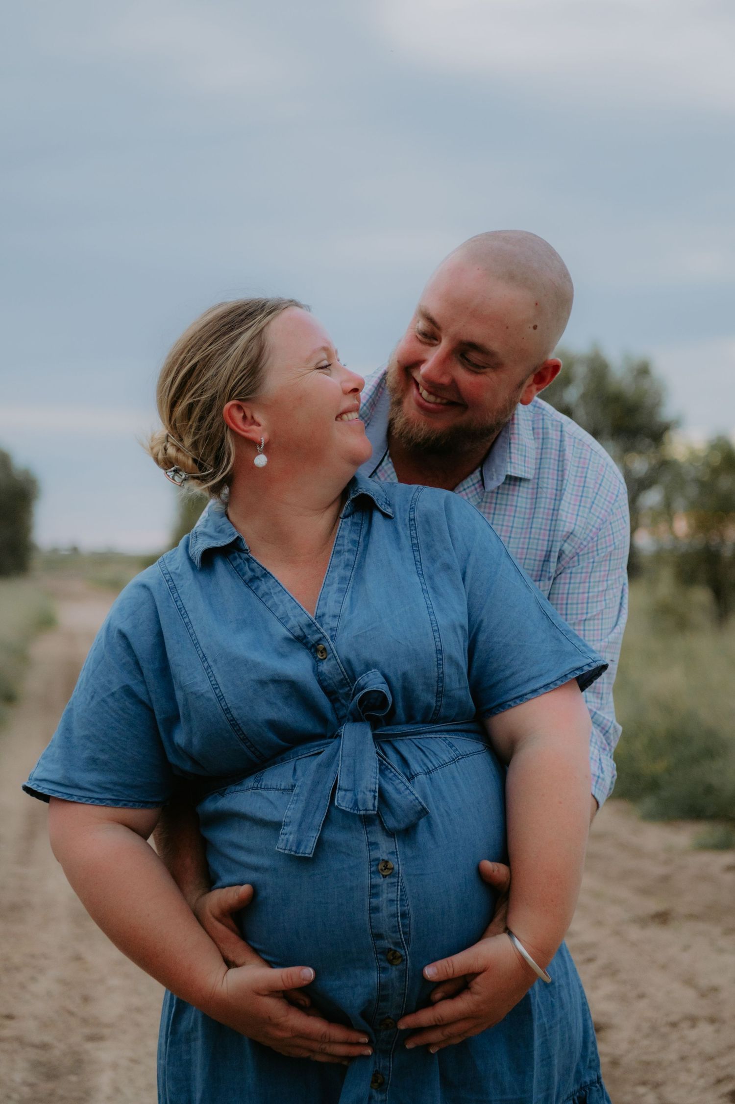 Smiling pregnant couple embracing outdoors on a dirt path, woman in blue denim dress cradling baby bump.