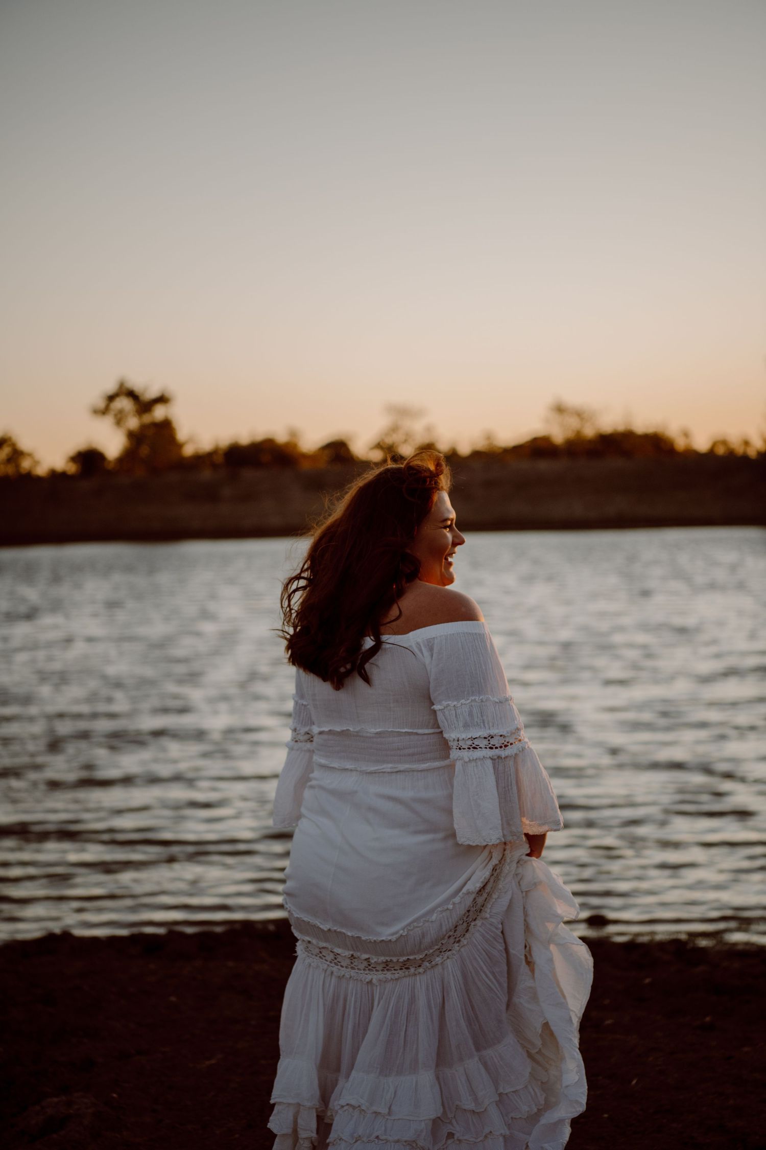 A silhouette of a woman in a flowing white dress standing by a lake at sunset.
