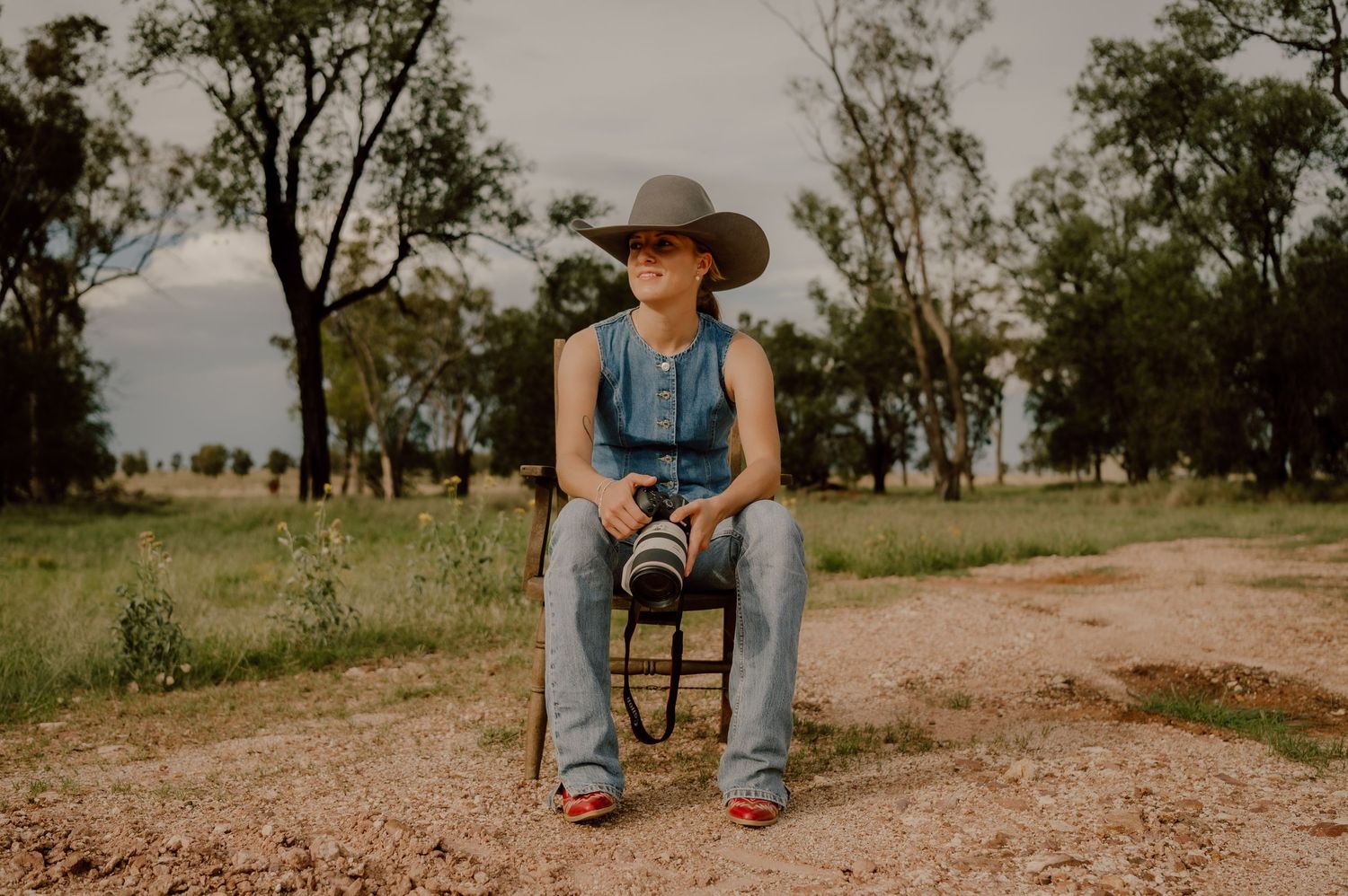 A person in a cowboy hat and denim sits on a dirt road in a rural landscape at sunset.