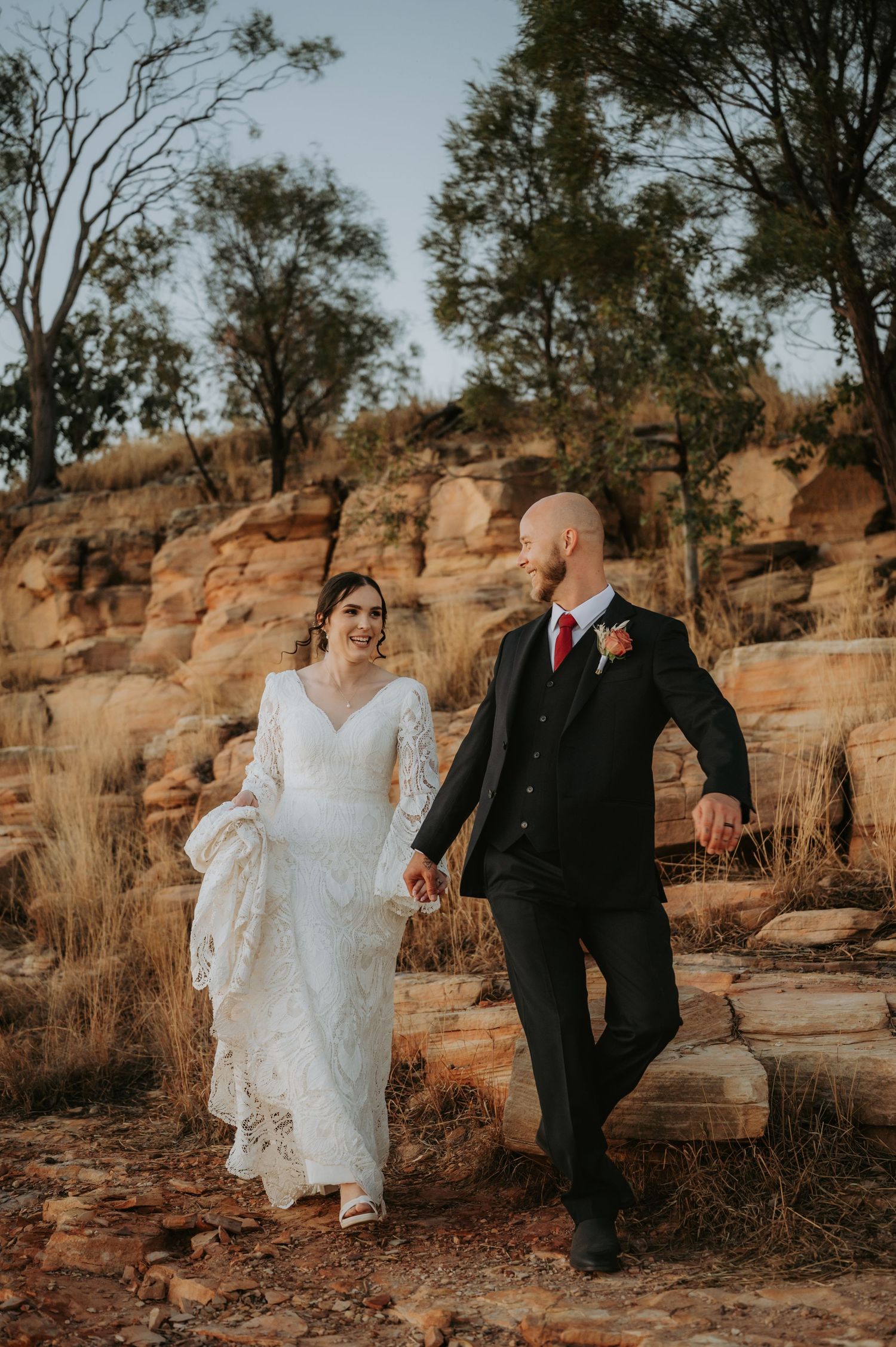 Bride and groom walk hand-in-hand among red rock formations in an Australian outback wedding setting.