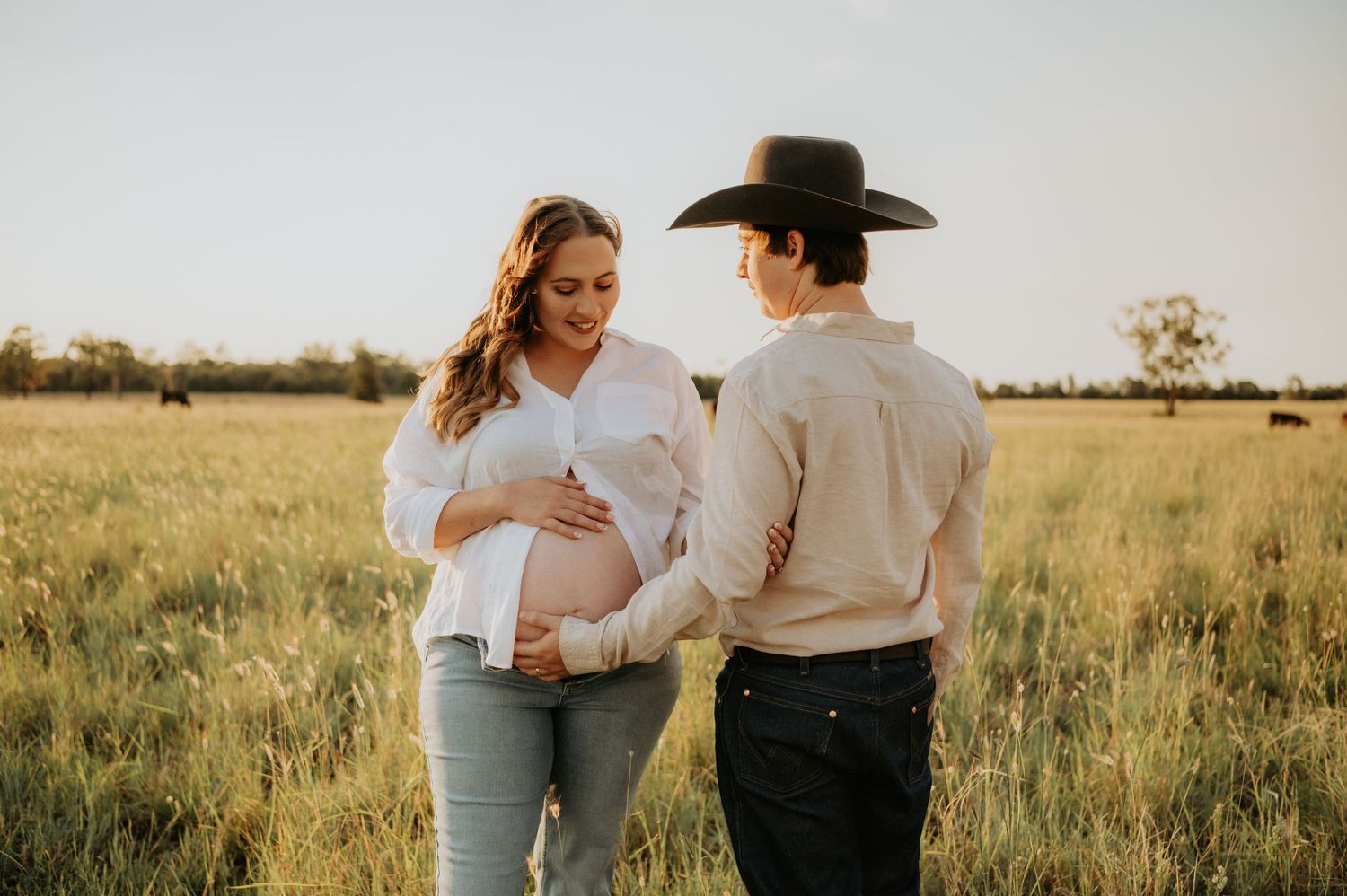 Pregnant couple sharing a tender moment in a golden sunlit field, man wearing a cowboy hat touching her baby bump.