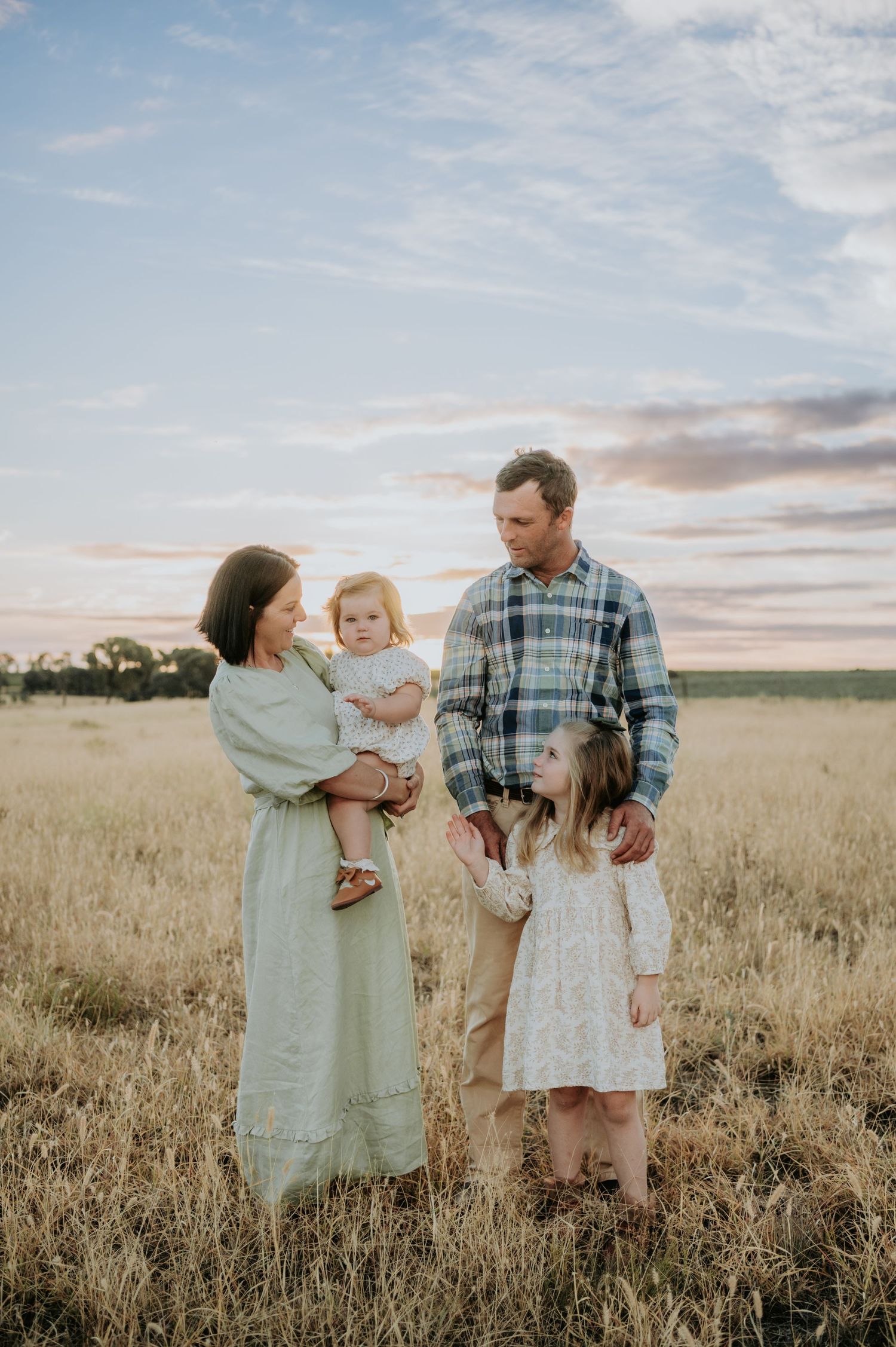 Family of four posing in golden field at sunset, mother holding toddler, father standing beside older daughter in white dress.