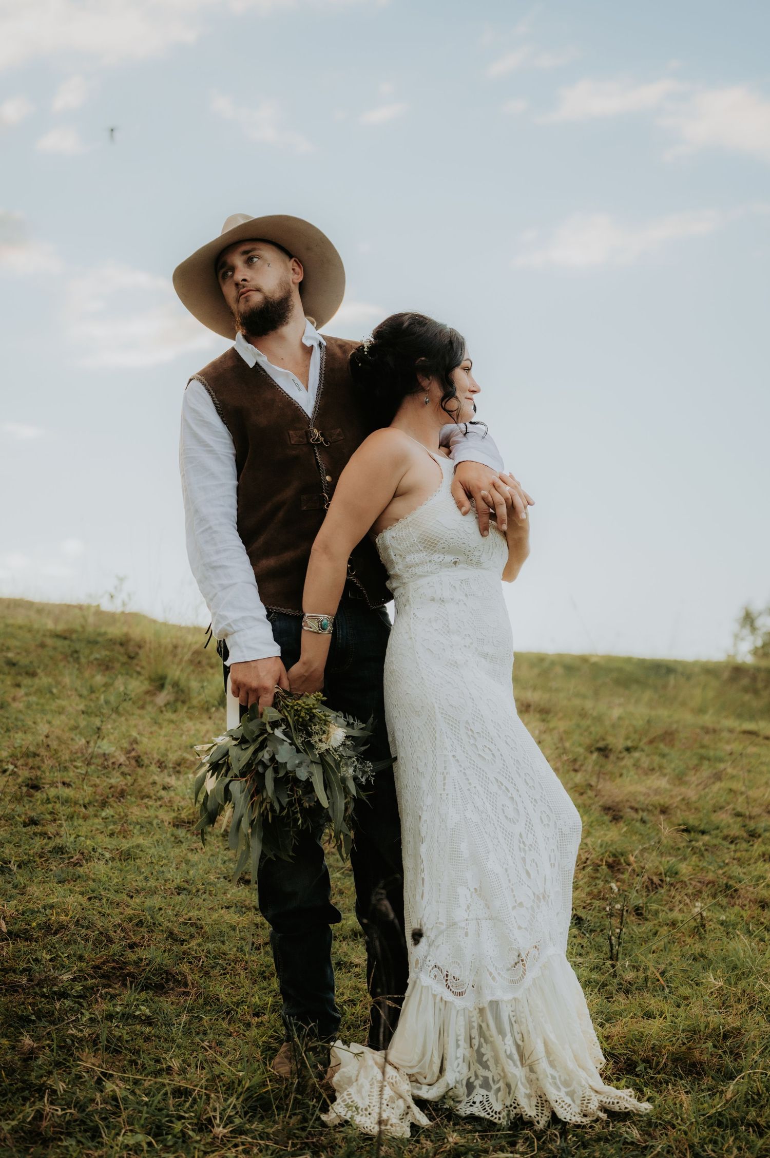 Rustic outdoor wedding couple, groom in brown vest and wide-brim hat holding bouquet, bride in lace gown on green hillside.